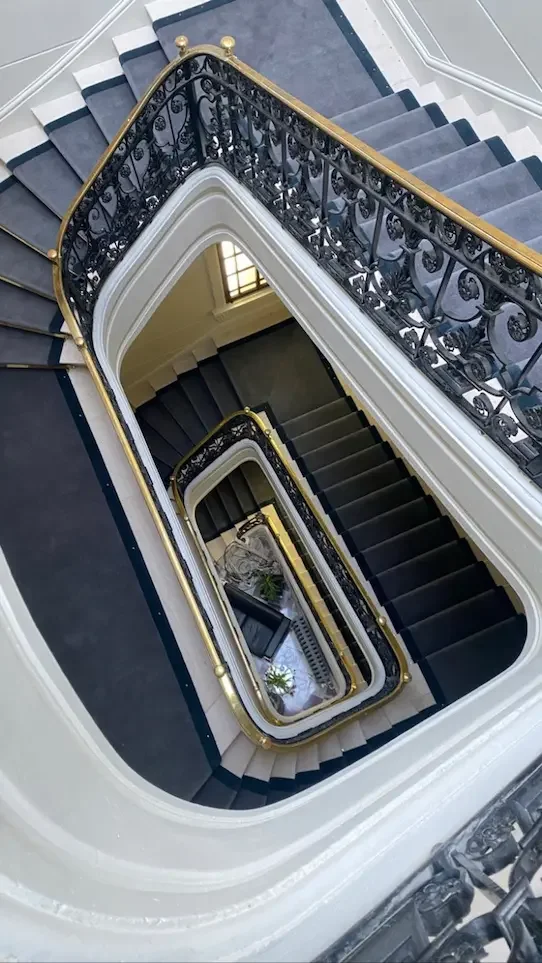 Vue d'en haut d'un escalier en spirale avec des marches noir et blanc, une rambarde en fer ornemental noir avec des détails en or, dans un bâtiment élégant.