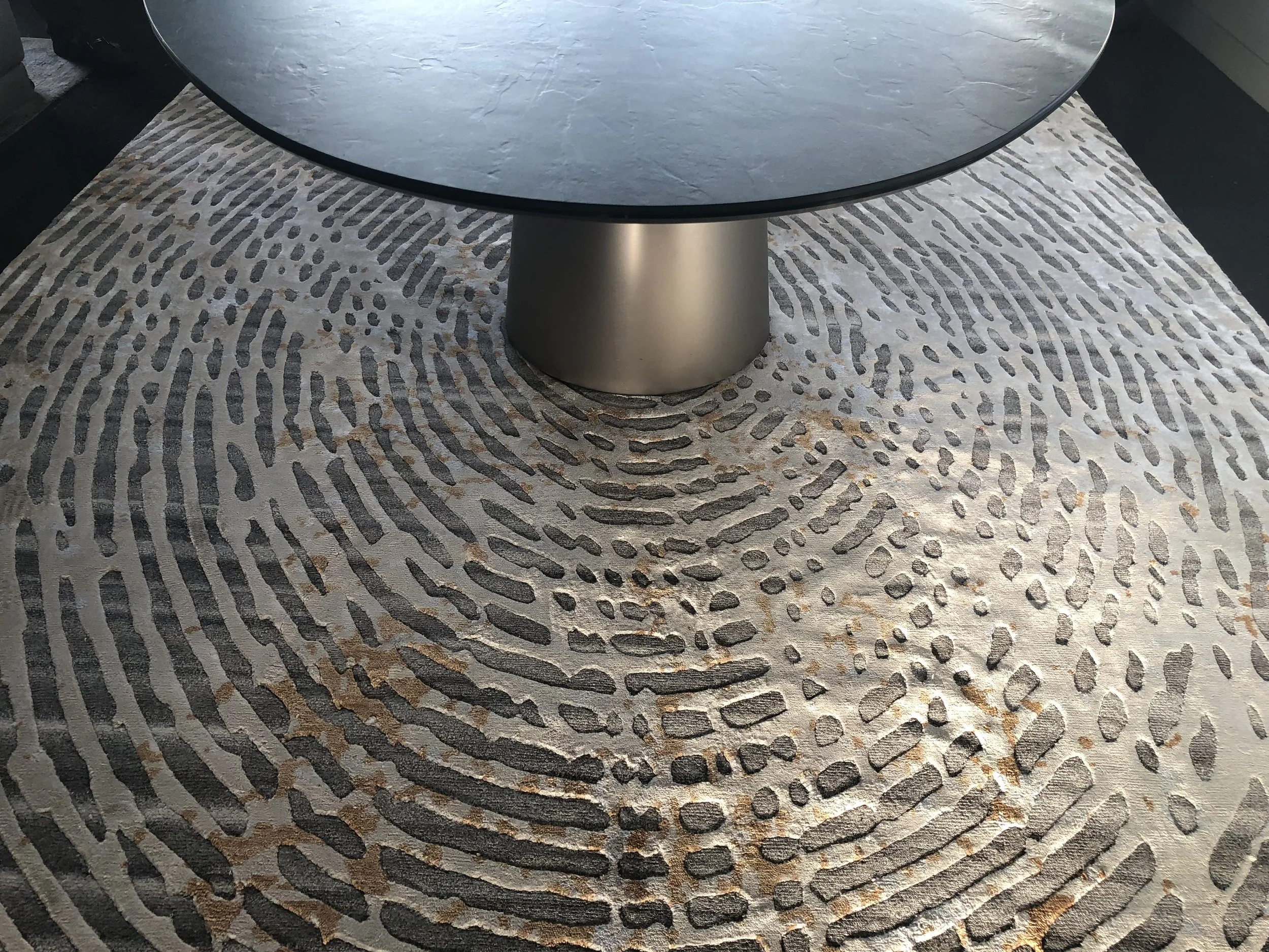 Table ronde en pierre avec une base métallique ronde posée sur un tapis décoratif aux motifs abstraits en gris et beige.