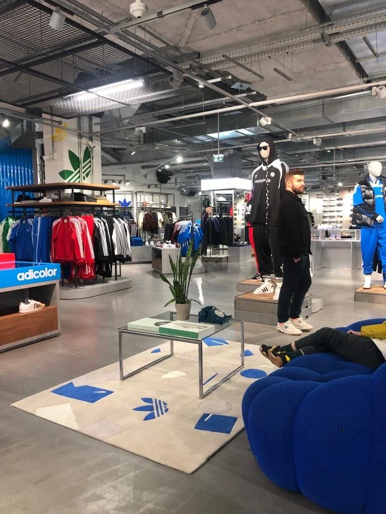 Intérieur d'un magasin de vêtements de sport avec des mannequins, des rayons de vêtements, et un espace de détente avec un tapis, un fauteuil bleu et une table
