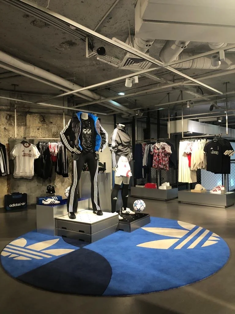 Mannequins habillés en vêtements de sport dans une boutique Adidas, avec des vêtements et des accessoires sur le fond, et un tapis rond bleu avec le logo Adidas.