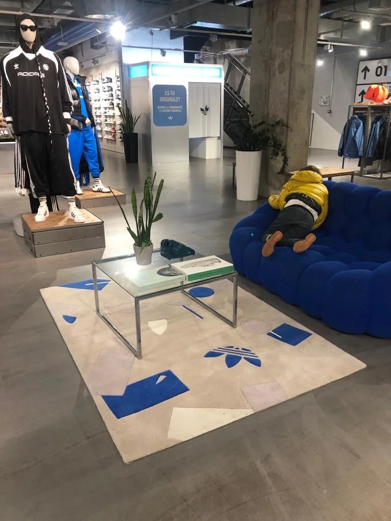 Mannequins vêtus de vêtements de sport Adidas dans un magasin, canapé bleu avec enfant allongé, tapis beige avec motifs bleus, table en verre avec plantes et livres.