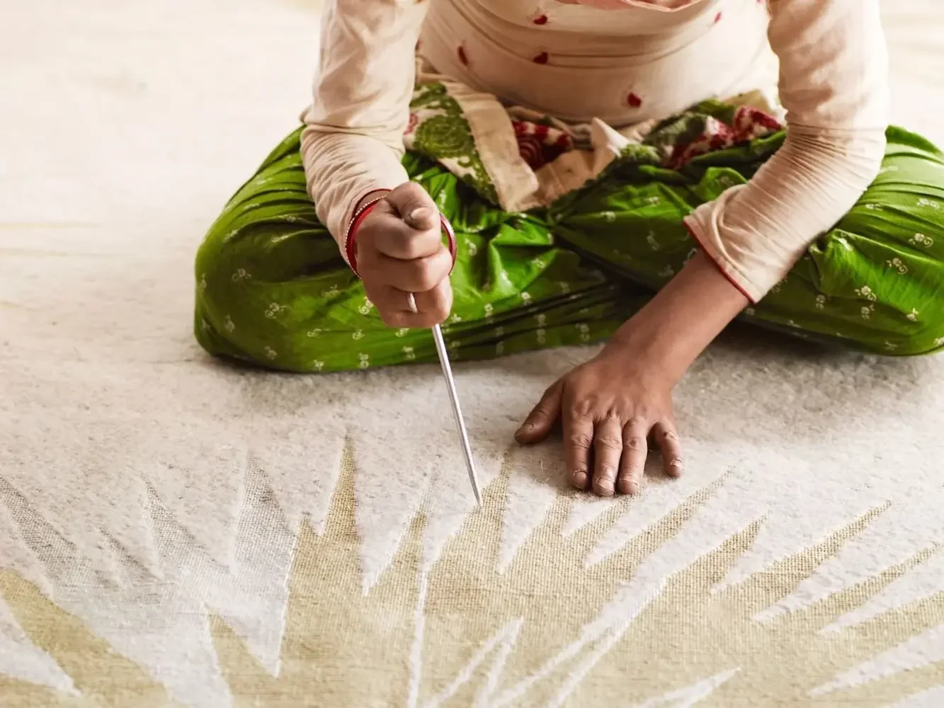 A rug depicting a person sitting cross-legged on it, using a tool to create a sun pattern painted on the surface.