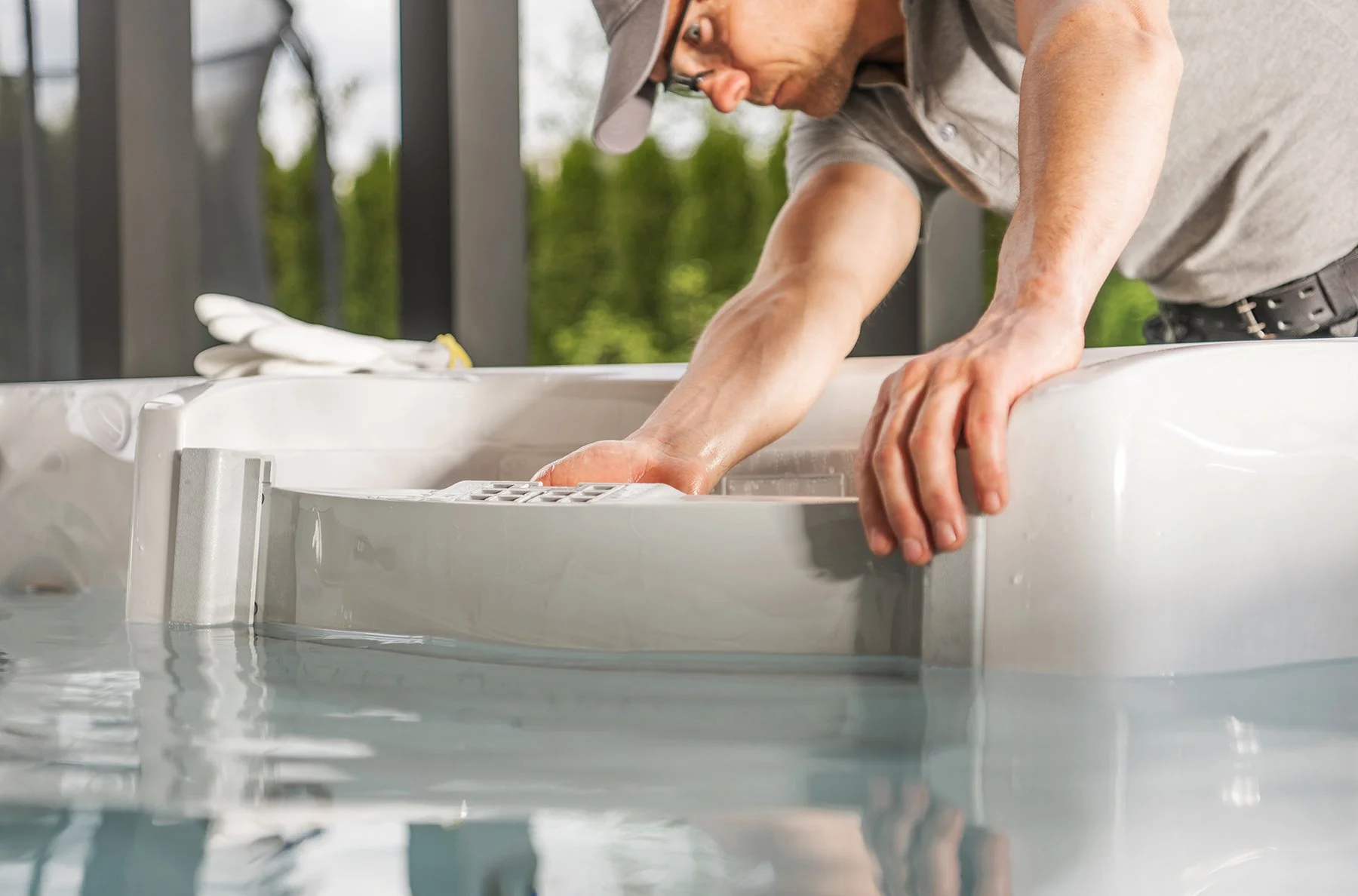 Man cleaning a hot tub while leaning over the edge, wearing a cap and glasses, with a background of trees.