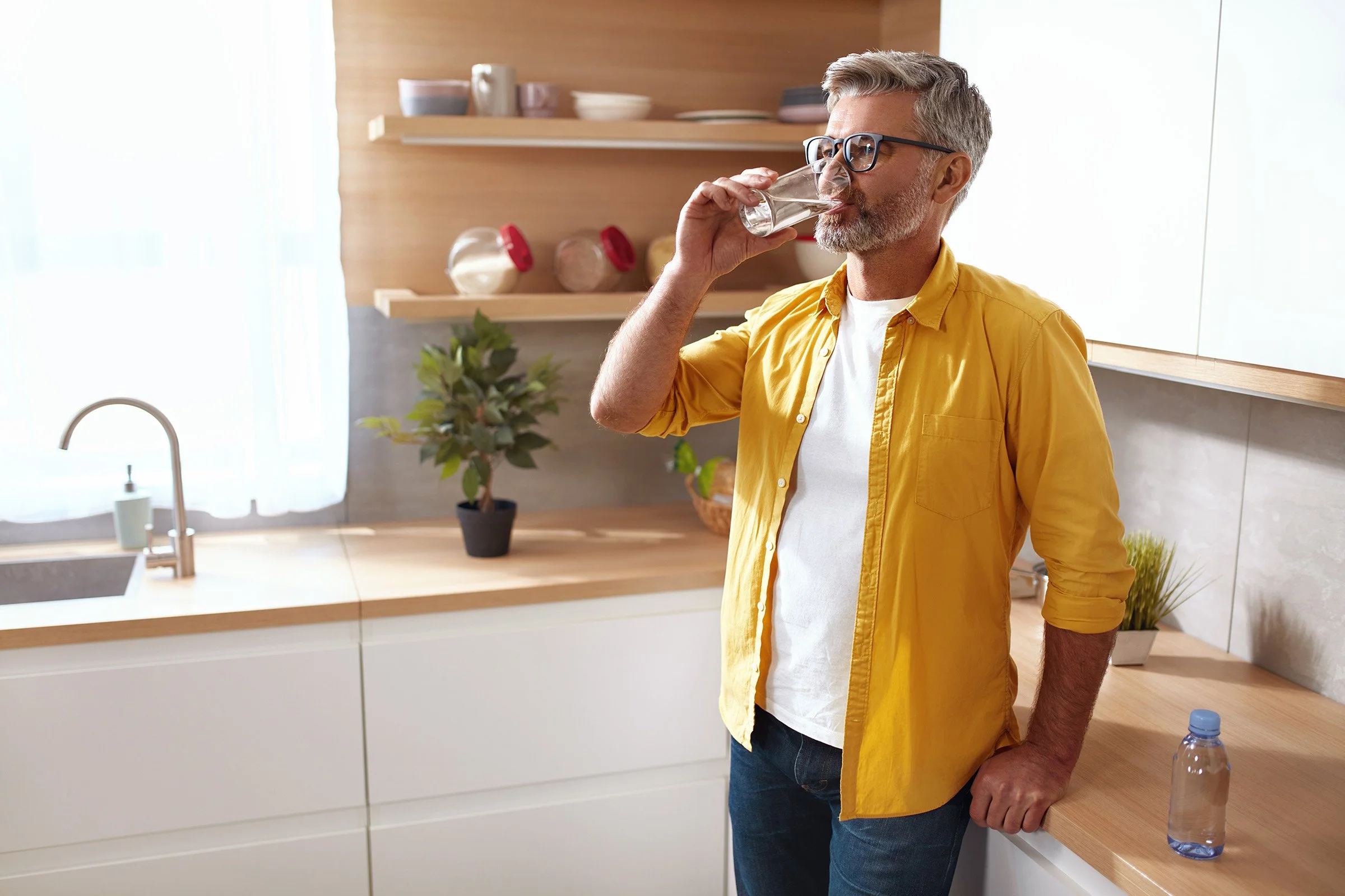 Man with gray hair, glasses, wearing a yellow shirt, drinking water in a modern kitchen with wooden shelves, potted plants, and sunlight.