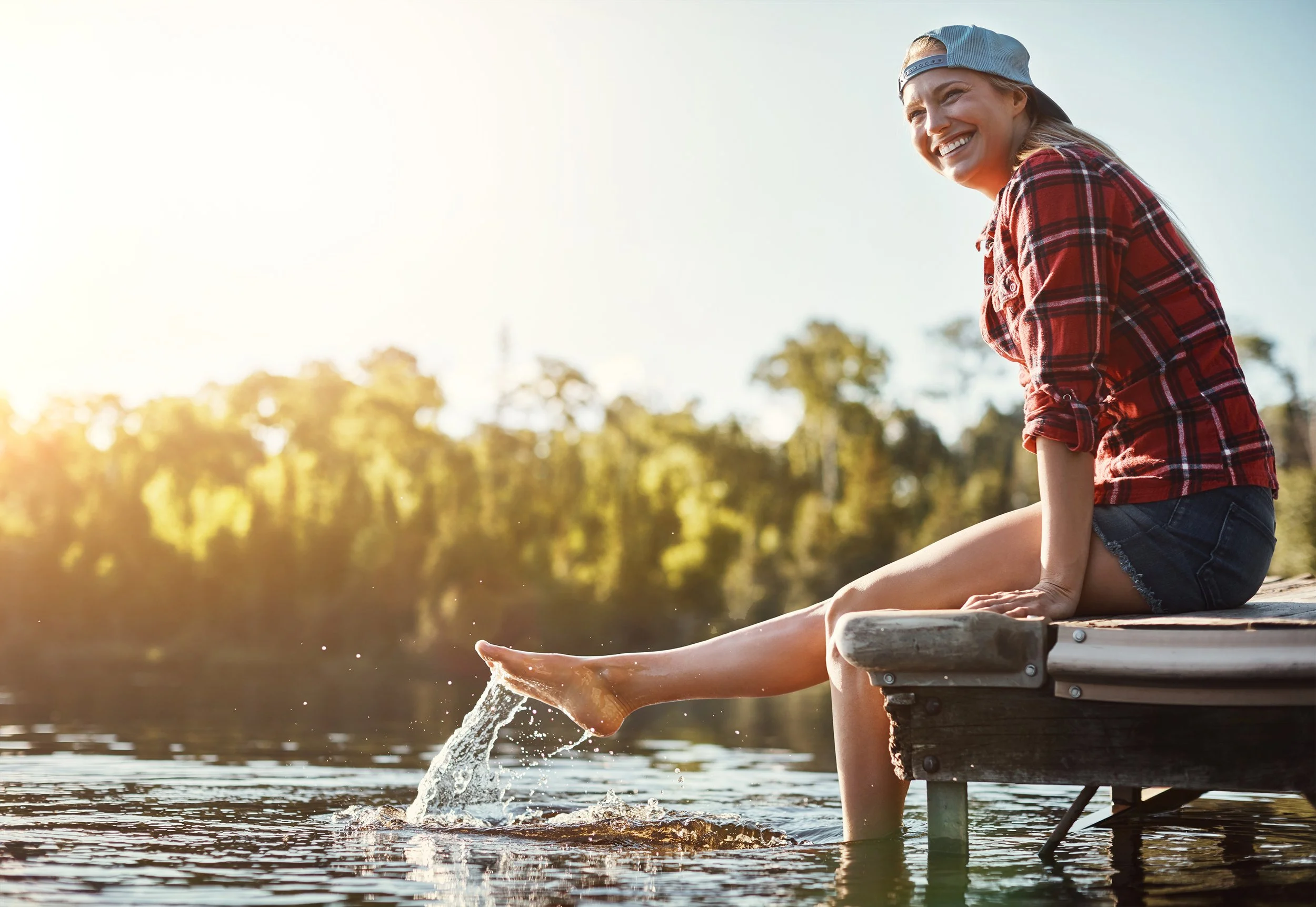 A young woman sits on a dock by a lake, dipping her feet into the water, smiling, wearing a baseball cap backward, a red plaid shirt, and denim shorts in late afternoon sunlight.