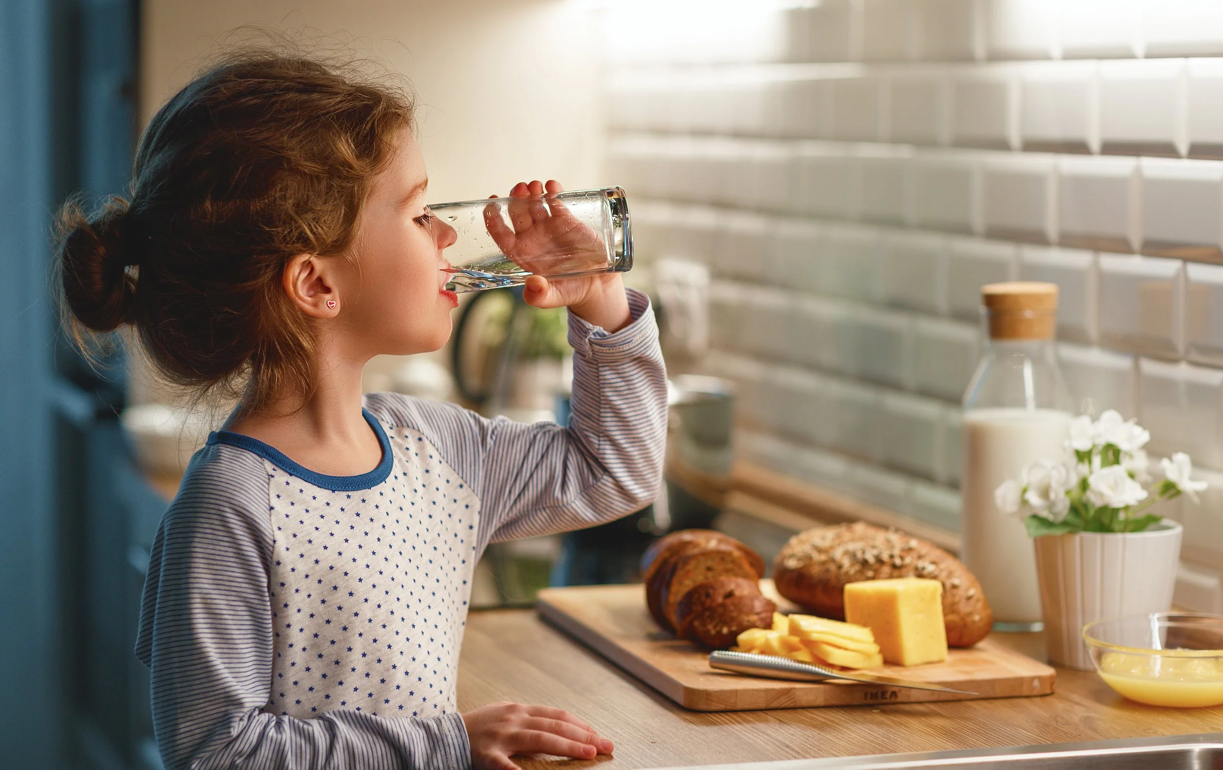 A young girl with curly hair and heart-shaped earrings drinks a glass of water in a kitchen, with baked bread, cheese, milk, and flowers on the counter.