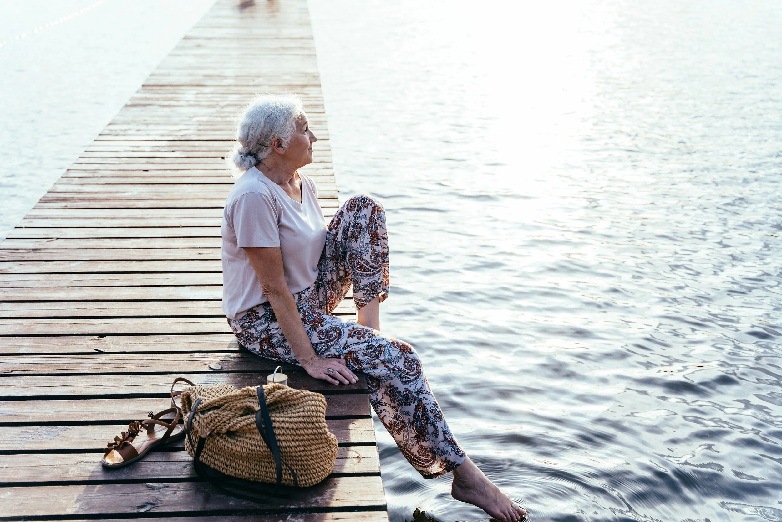 An elderly woman with gray hair sitting on a wooden dock by the water during sunset, with a woven bag and sandals beside her.