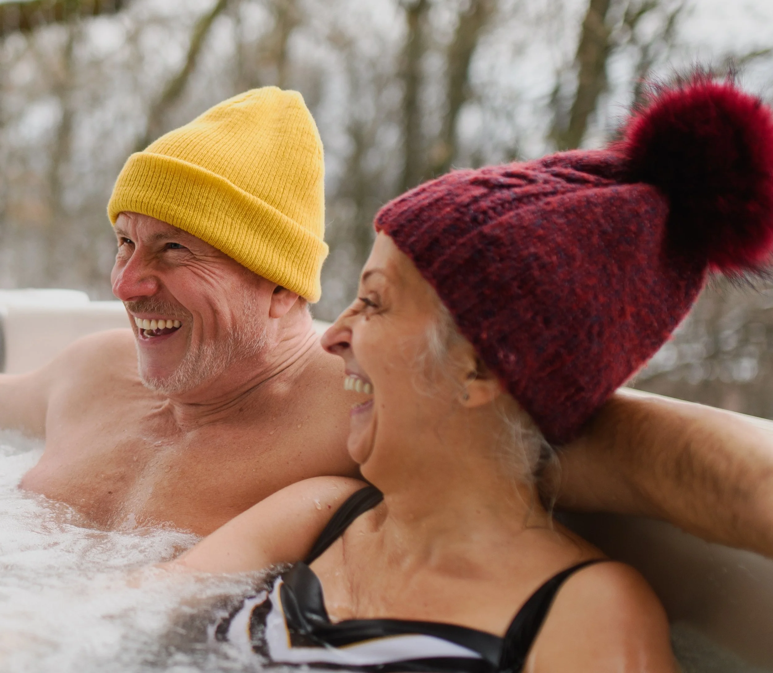 An elderly man and woman laughing and enjoying time together outdoors in a hot tub, wearing colorful knit hats.