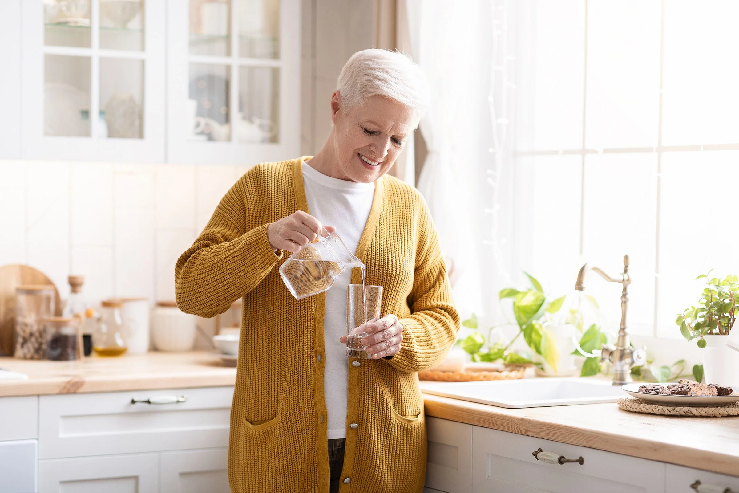 An elderly woman is pouring water from a pitcher into a glass in a bright kitchen with white cabinets and a window.