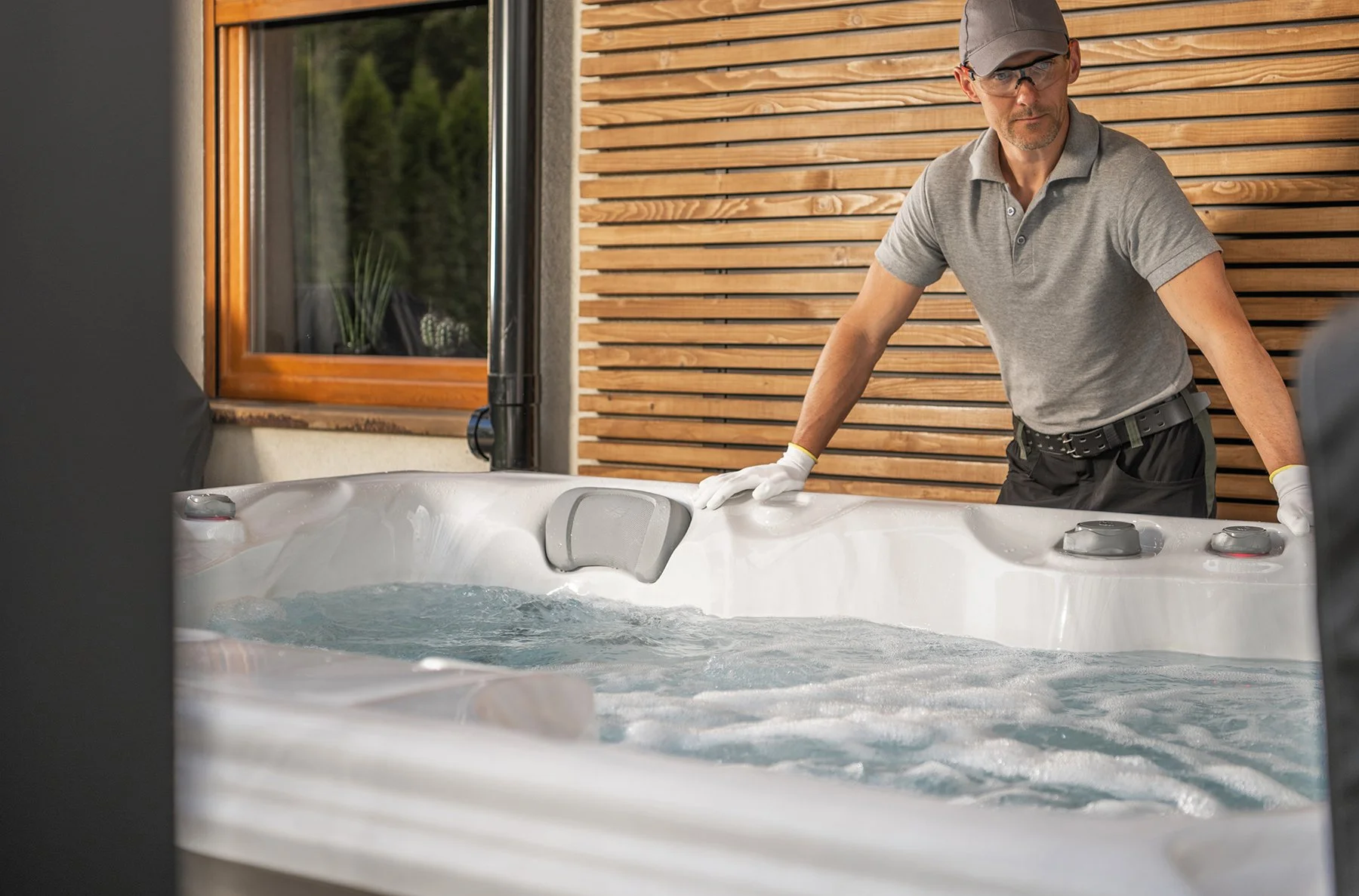 A man wearing a gray polo shirt, glasses, and a baseball cap is cleaning or inspecting a large outdoor hot tub or jacuzzi on a patio with wooden paneling. The hot tub has bubbling water and control panels.