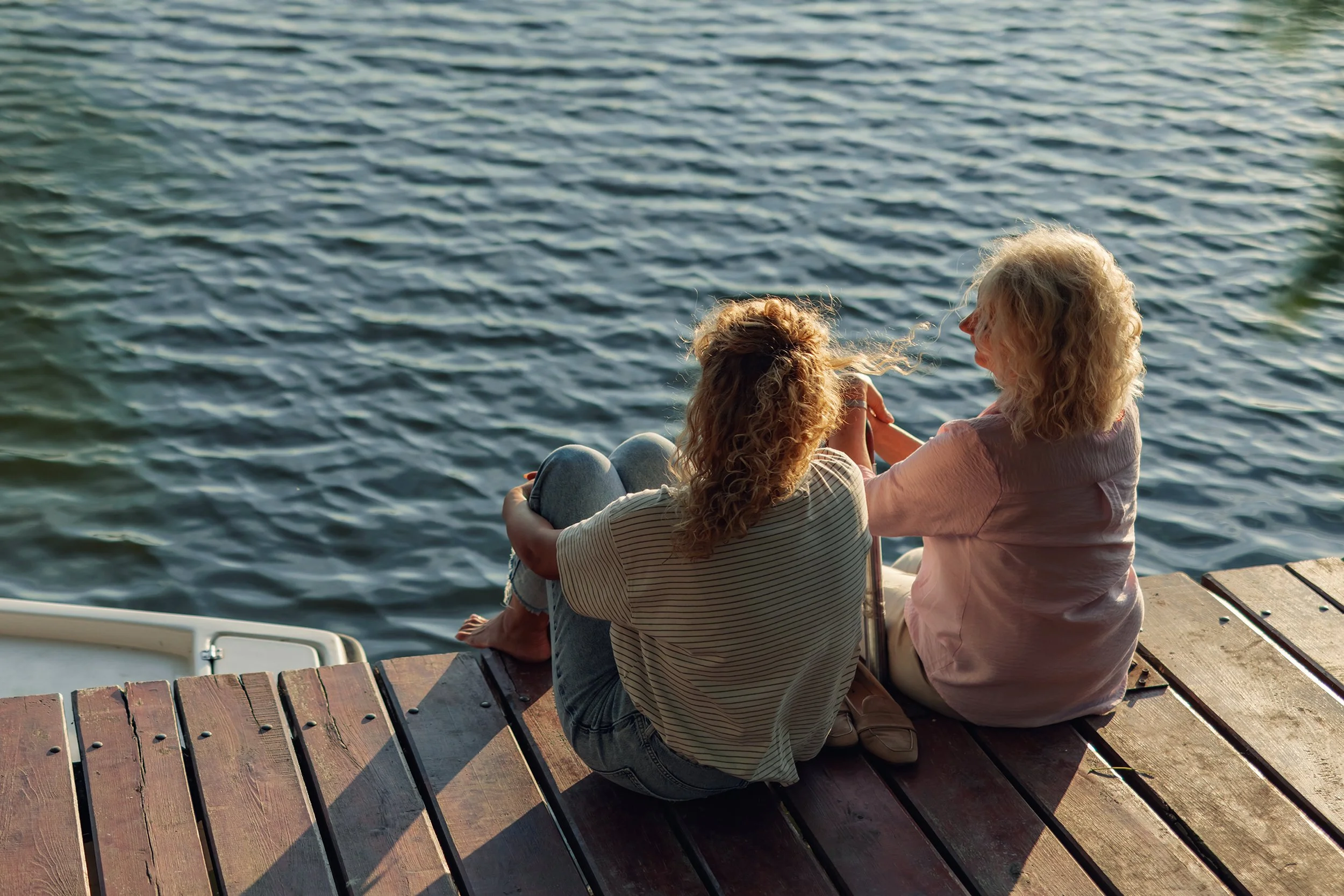 Two women sitting on a wooden dock by the water, sharing a moment and holding hands, with water in the background.