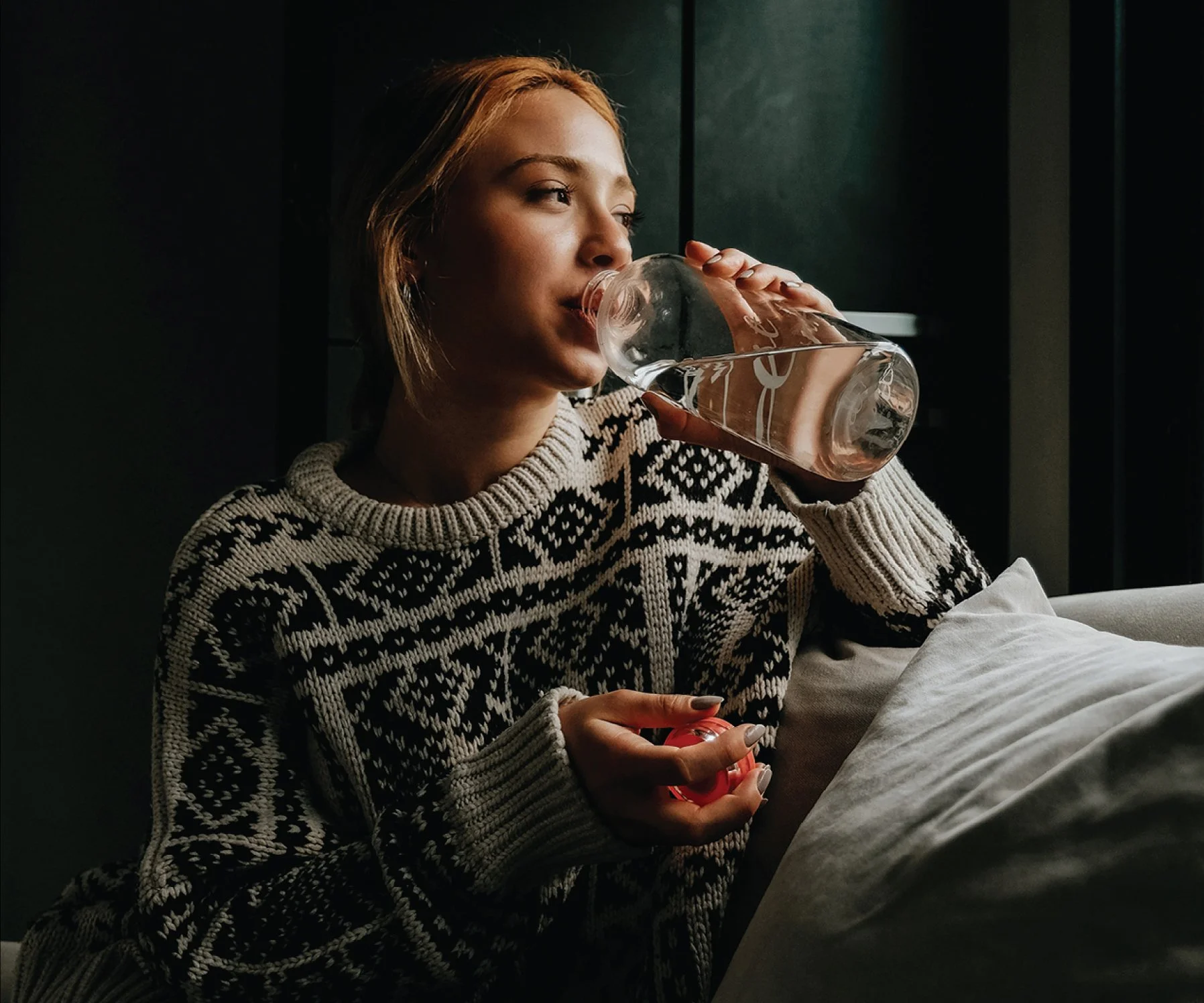 A woman with shoulder-length hair drinking water from a glass while sitting on a sofa, wearing a patterned winter sweater.