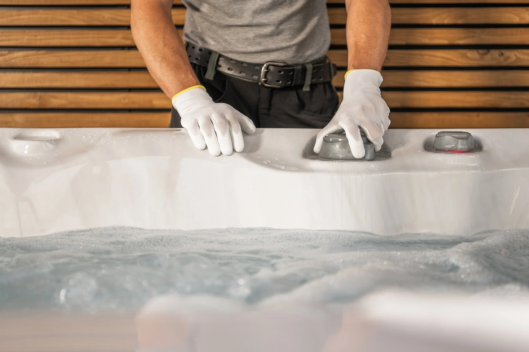 A person wearing gloves is cleaning or polishing a white bathtub with a cloth or sponge in a modern bathroom with wooden paneling.