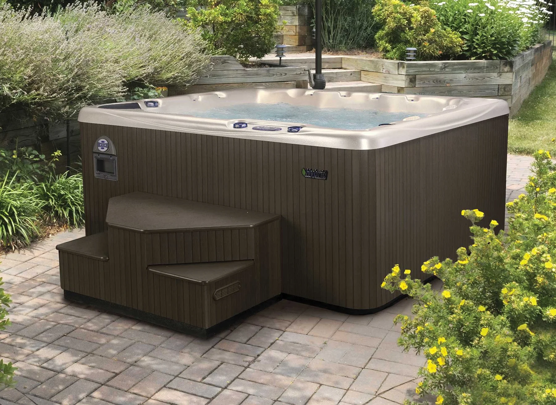 A hot tub with brown exterior panels and beige interior, with water filled inside, situated on a paved outdoor area surrounded by green bushes and yellow flowering plants.