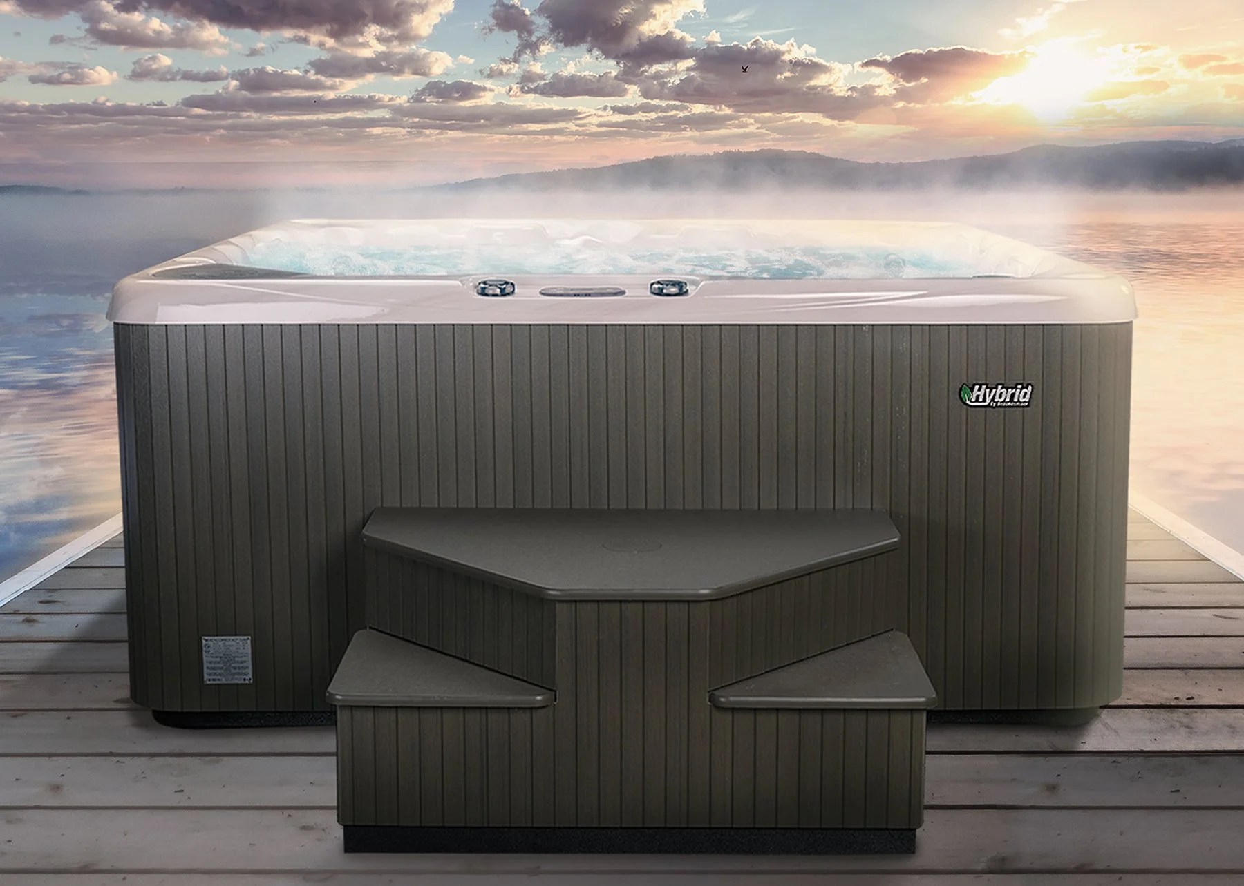 A hot tub with a black exterior and a built-in step is placed on a wooden dock overlooking a calm lake during sunset, with clouds and mountains in the background.