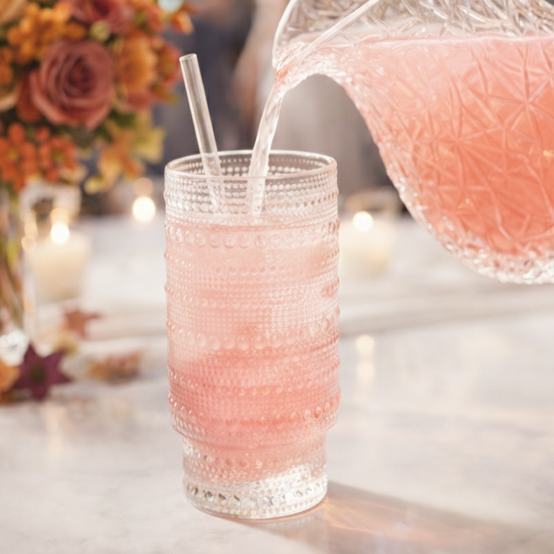 A glass with a textured pattern contains pink liquid and a straw, with pink liquid being poured from a patterned pitcher.