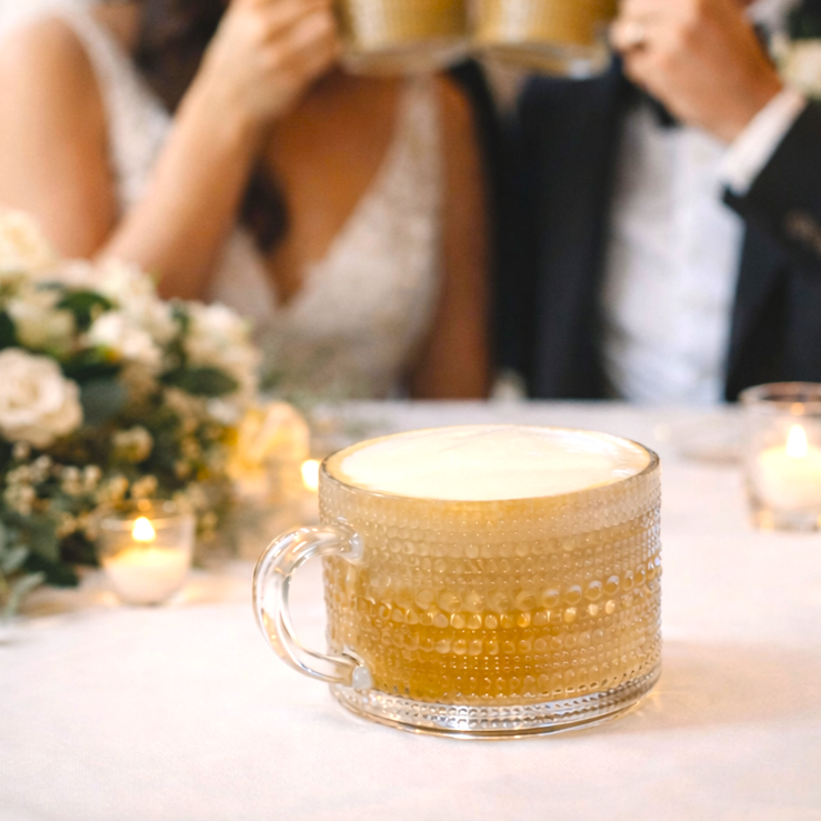 A man is feeding a woman a drink through a glass window at a cafe or restaurant. The woman is smiling and holding a glass, and they are holding hands on the table. The scene is warm and intimate.