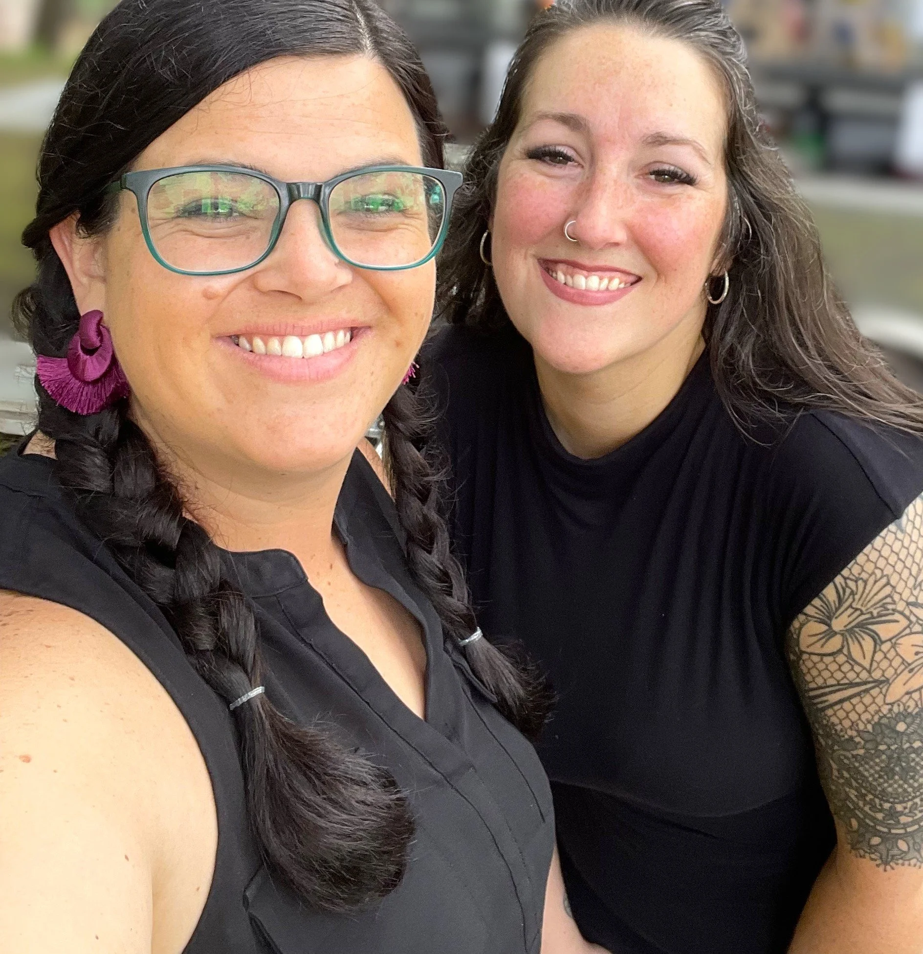 Two women smiling outdoors, one with glasses and braids, the other with a nose ring and tattooed arm, both wearing black tops.