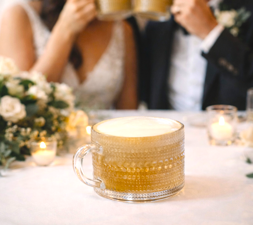 A latte sitting on a white table, with a blurred background of a wedding reception table, flowers, and candles.