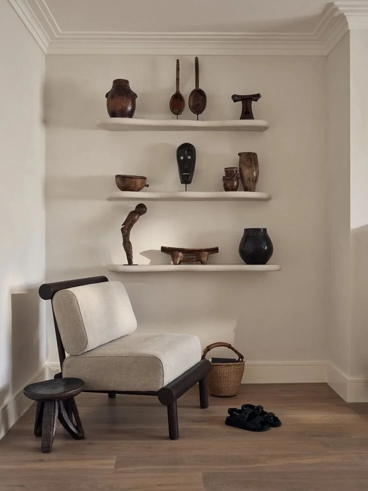 A white wall with three floating shelves displaying various wooden and ceramic decorative items, a beige cushioned chair with dark wooden frame, a small rustic stool, a woven basket, and a pair of black shoes on the wooden floor.