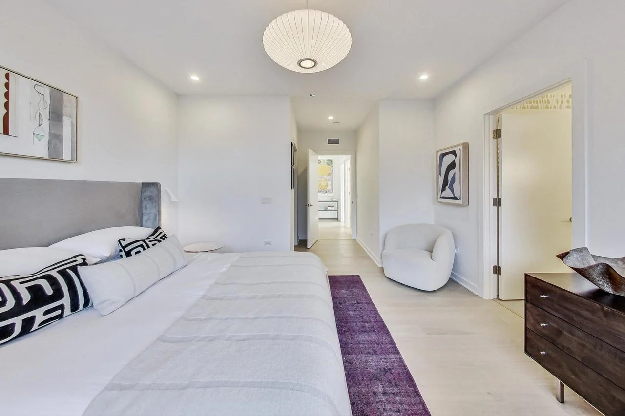 A modern bedroom with a large bed, white bedding, decorative pillows, and wall art. There is a white armchair, a dark wooden dresser, and a purple rug on light hardwood floors. Light fixtures include a ceiling lantern and recessed lighting.
