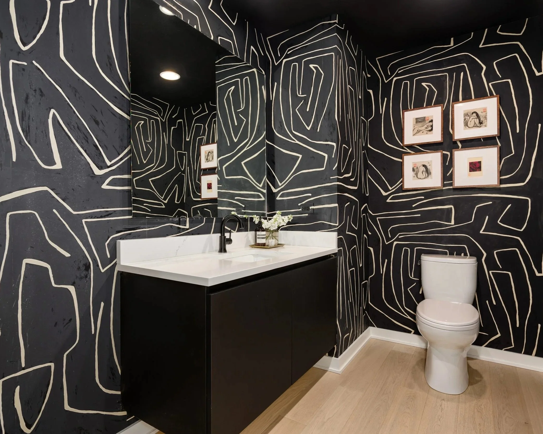 Modern bathroom with black and white abstract wall design, white toilet, black countertop with sink, large mirror, framed artwork on wall, wooden floor, and small vase with flowers.