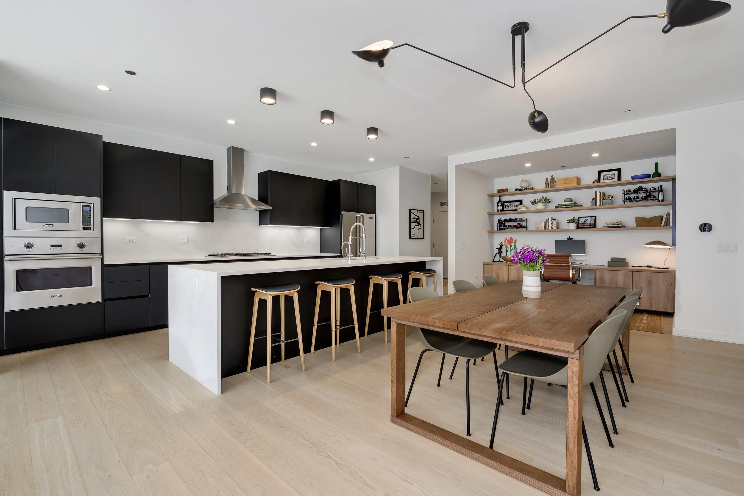 Modern open-concept kitchen and dining area with black cabinets, white island, wooden dining table, and built-in bookshelf.