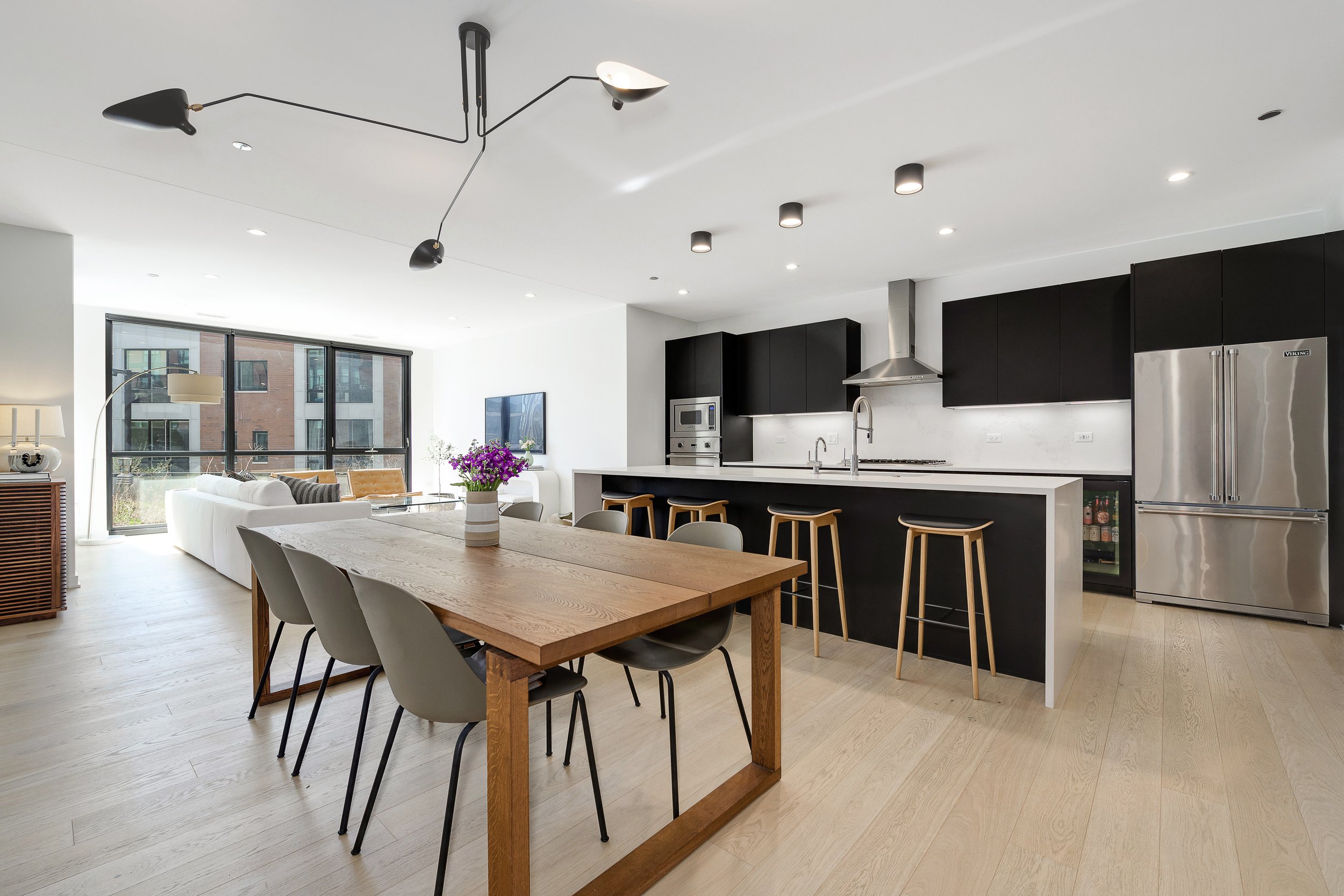 Open-concept modern kitchen and dining area with black cabinets, stainless steel appliances, wooden dining table, and bar stools.