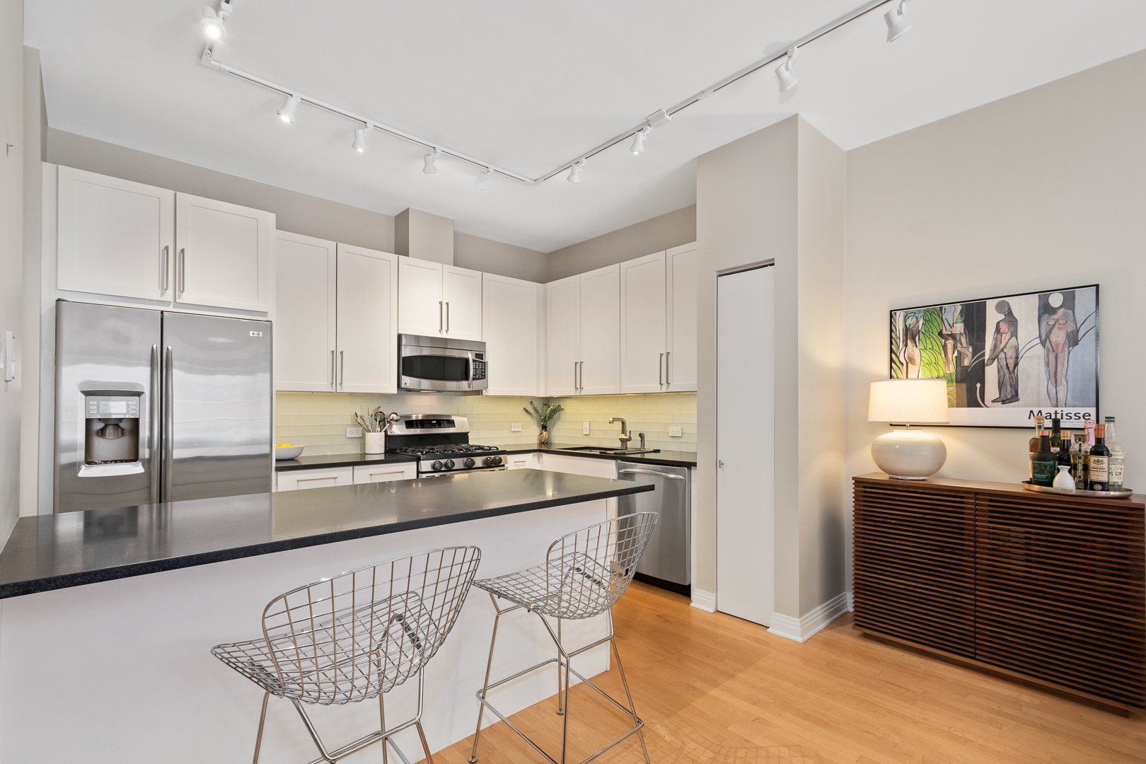 Modern kitchen with white cabinets, stainless steel refrigerator, microwave, and stove, black countertop, light wood floor, and a wooden sideboard with a lamp and bottles of wine underneath a colorful Matisse artwork.