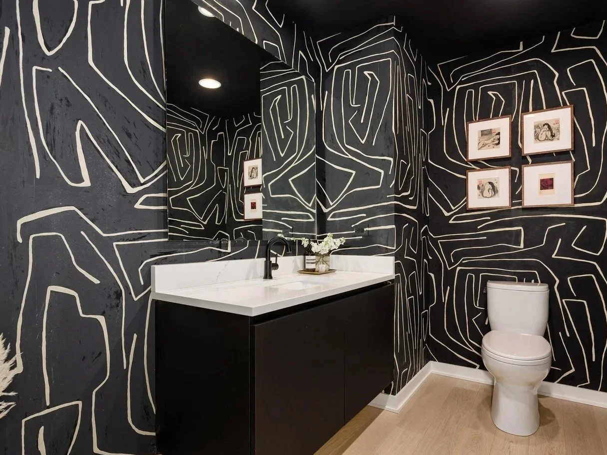 A modern bathroom with black walls decorated with white abstract line patterns. It features a white vanity with a black faucet, a large mirror, and framed artwork on the wall. There is a white toilet next to the vanity, and a small vase with white flowers on the countertop.