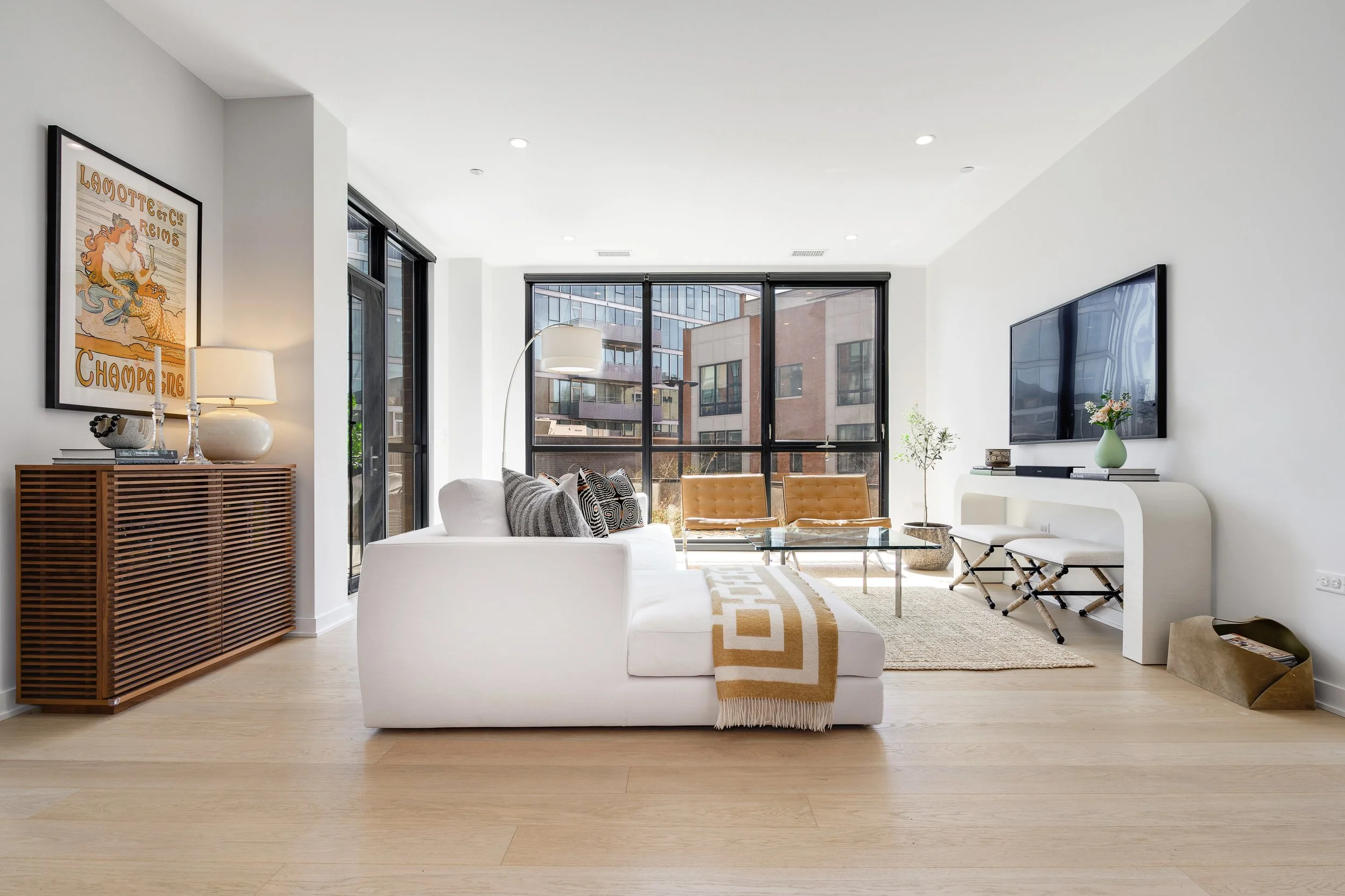 Modern living room with white sofa, two tan chairs, large windows, TV on white console, and decorative accents.