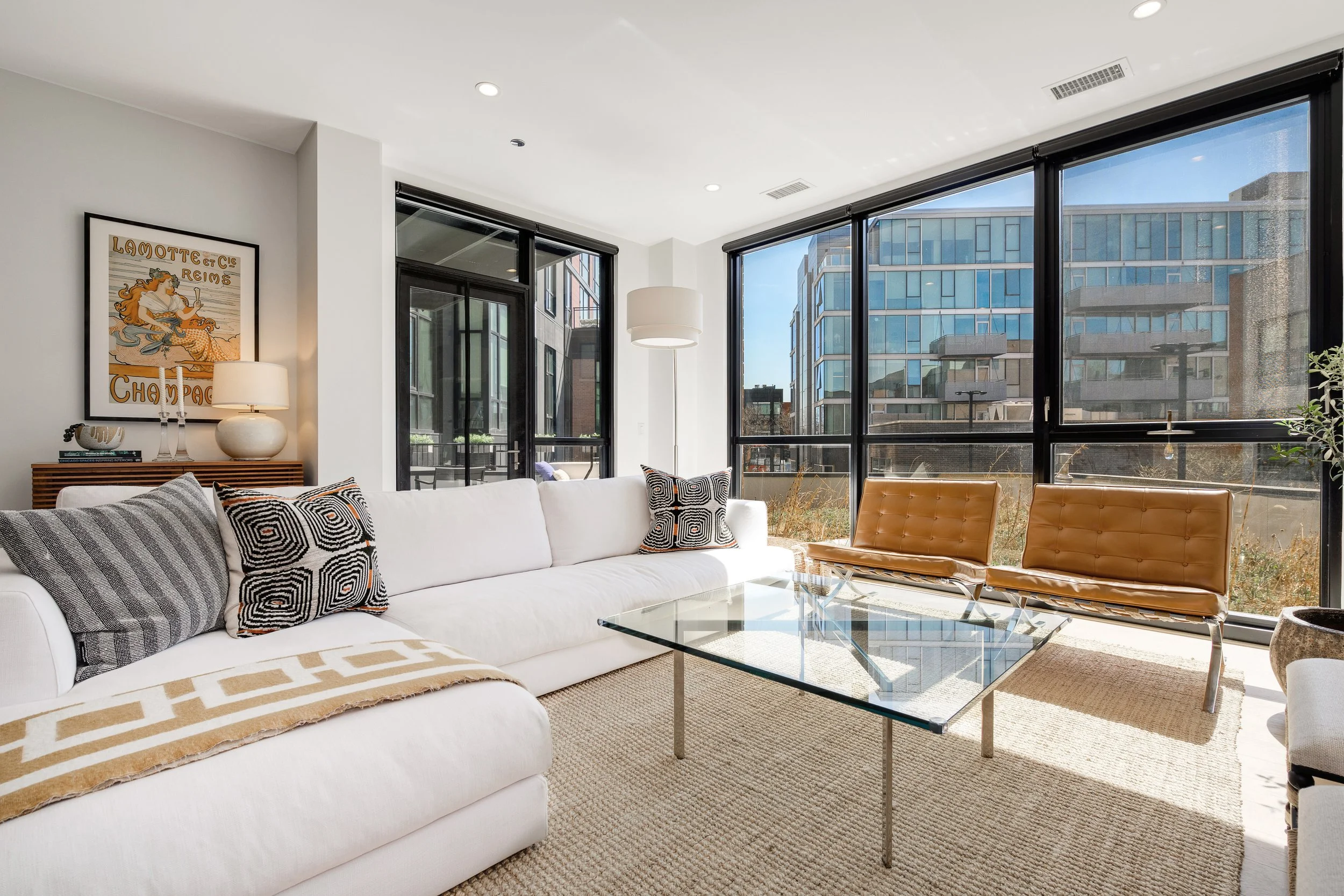Sunlit modern living room with white sofa, patterned cushions, glass coffee table, two tan Barcelona chairs, large windows, and urban view.