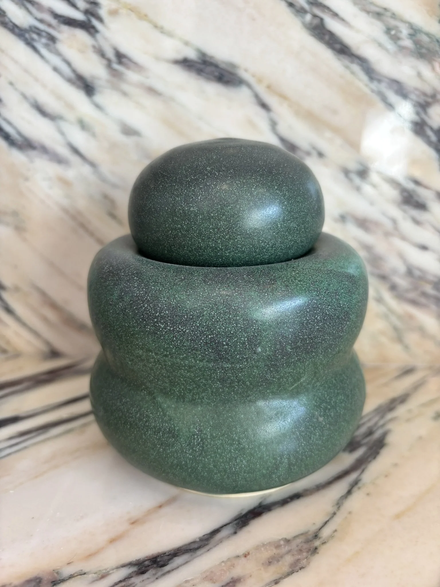 A stacked sculpture made of three smooth, rounded, green stones with speckled patterns, placed on a marble surface with a marble background.