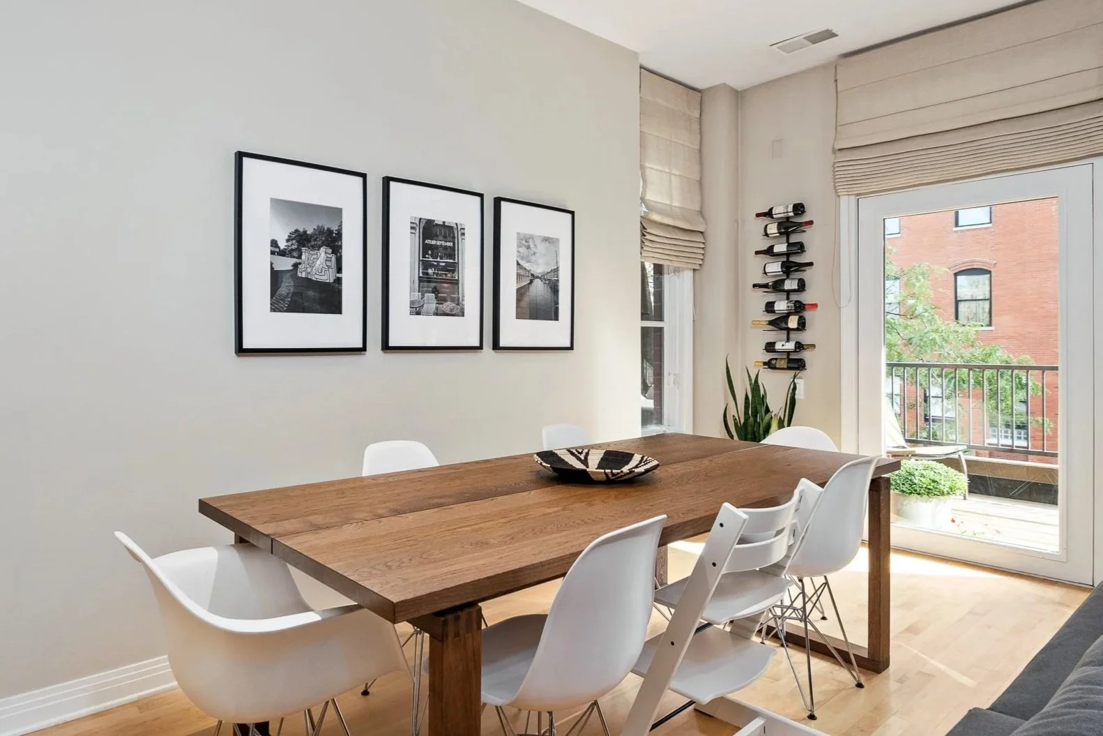 Dining room with a wooden table surrounded by white chairs, black and white framed photographs on the wall, a wine rack on the wall, a potted plant, and a balcony with a view of brick buildings.