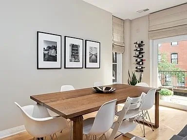 Modern dining room with wooden table, white chairs, framed black-and-white photos on the wall, and a balcony with plants outside.