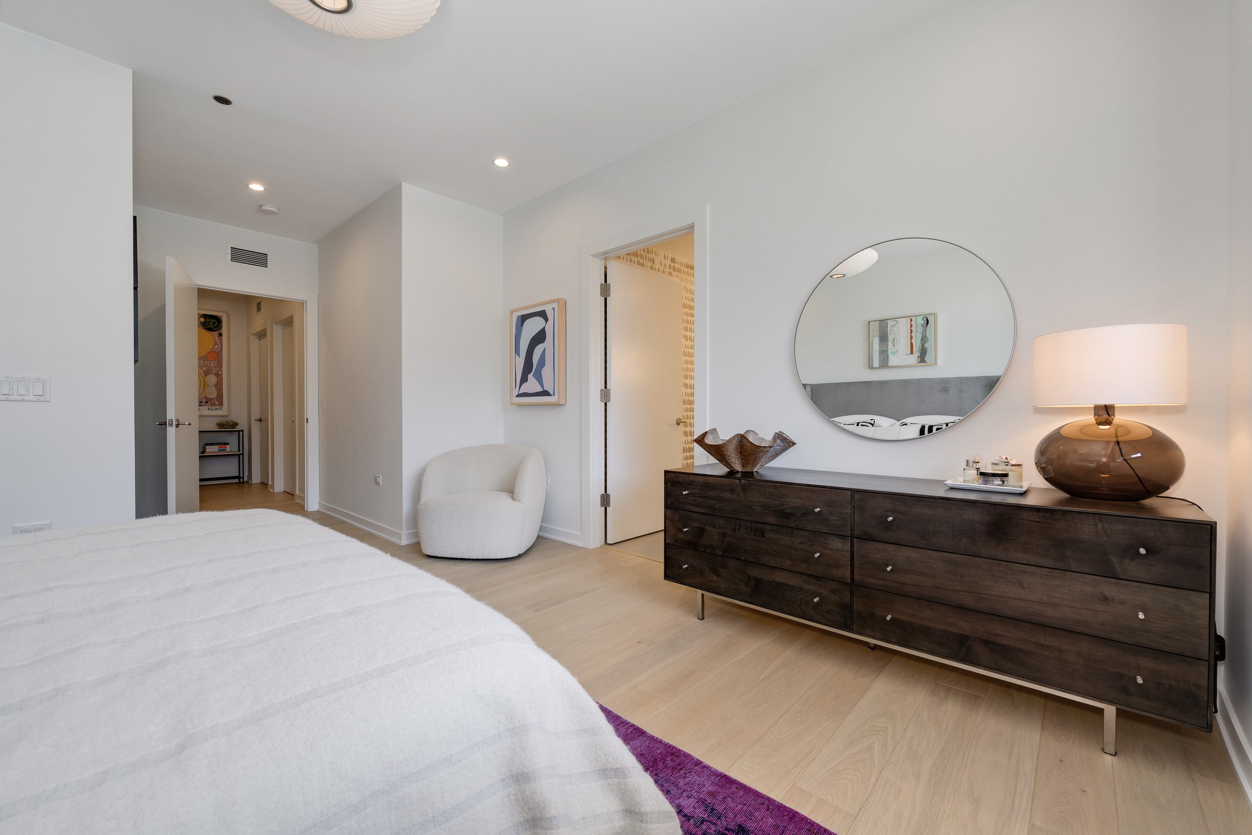 A modern bedroom featuring a white bed, a dark wood dresser with a mirror, a table lamp, a white upholstered chair, and art on the walls.
