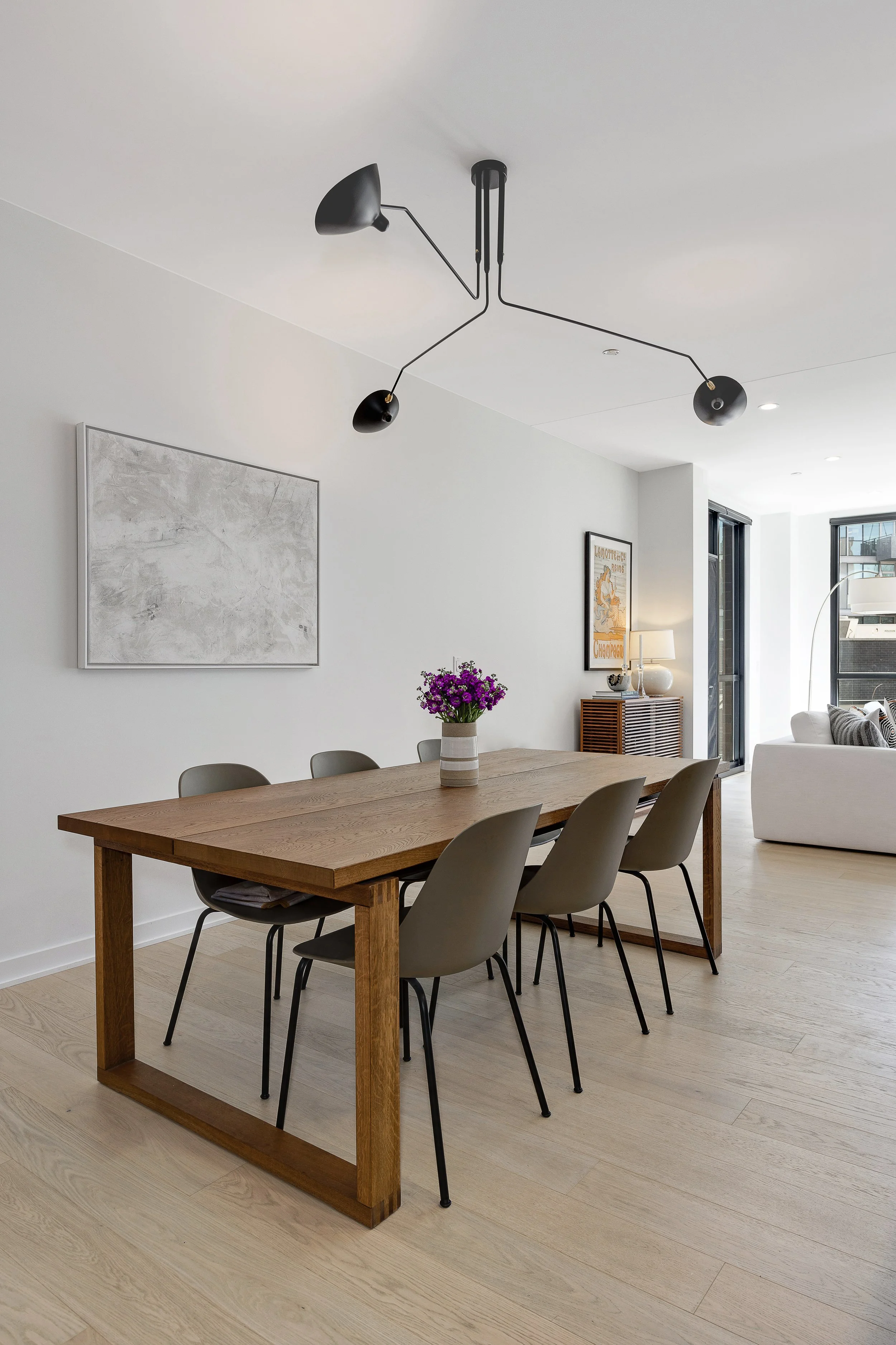 Modern dining room with a wooden table, six gray chairs, a vase with purple flowers, a hanging black light fixture, and a white sofa in the background.