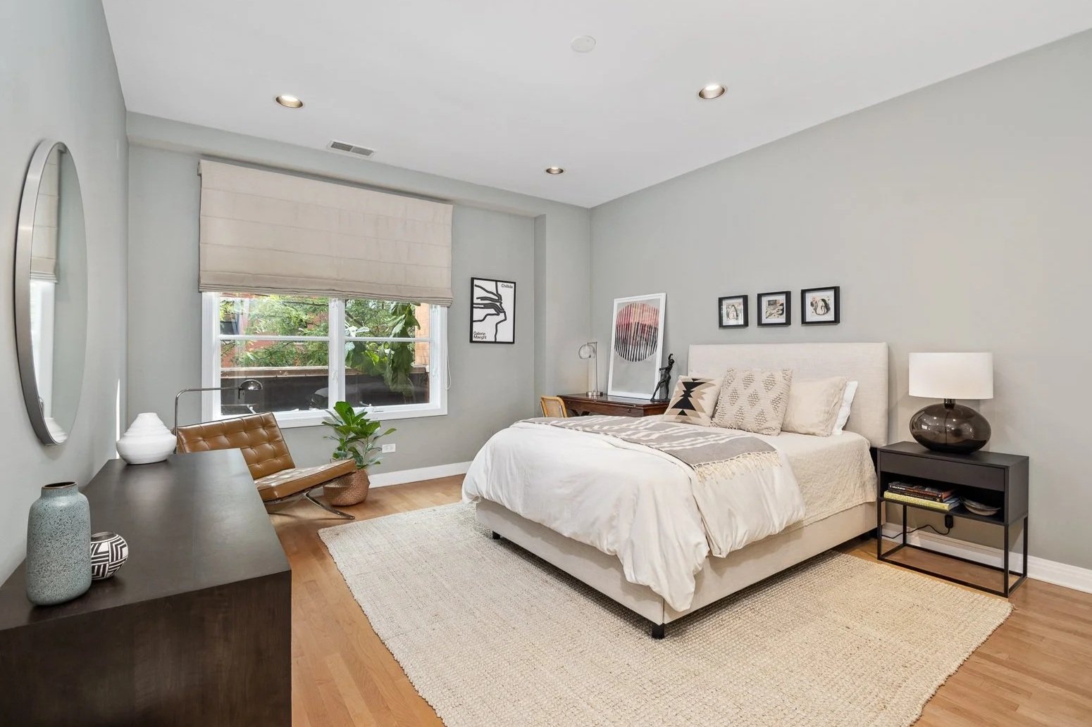 Modern bedroom with light gray walls, hardwood floors, a large bed with white linens, a black nightstand with a lamp, a black dresser with decorative vases, a chair near window with a plant, and framed artwork on the wall.