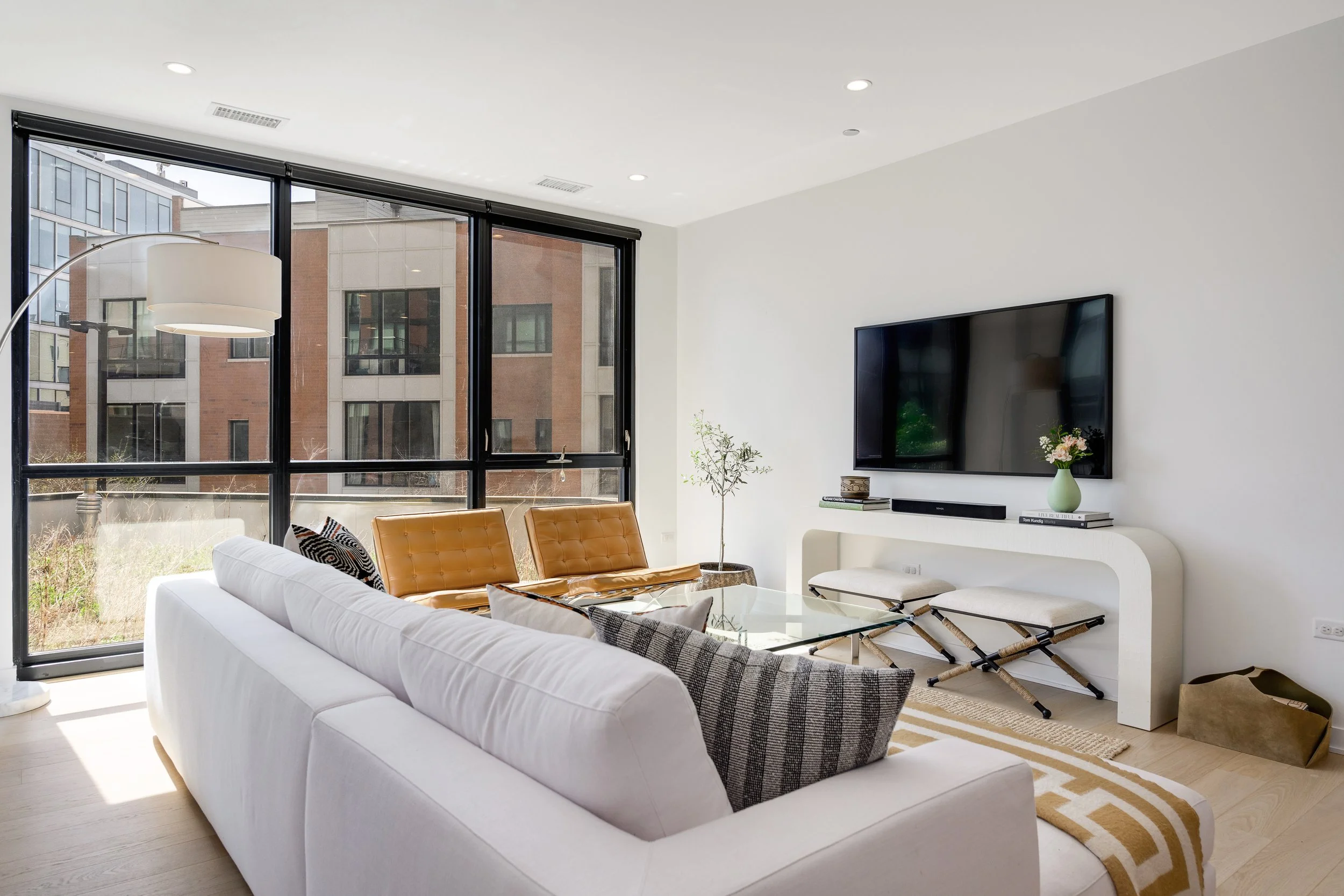 Modern living room with large windows, white sofa, glass coffee table, tan chairs, a TV mounted on the wall, a white console table, and a potted plant.