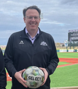 A man smiling at a baseball stadium holding a soccer ball.