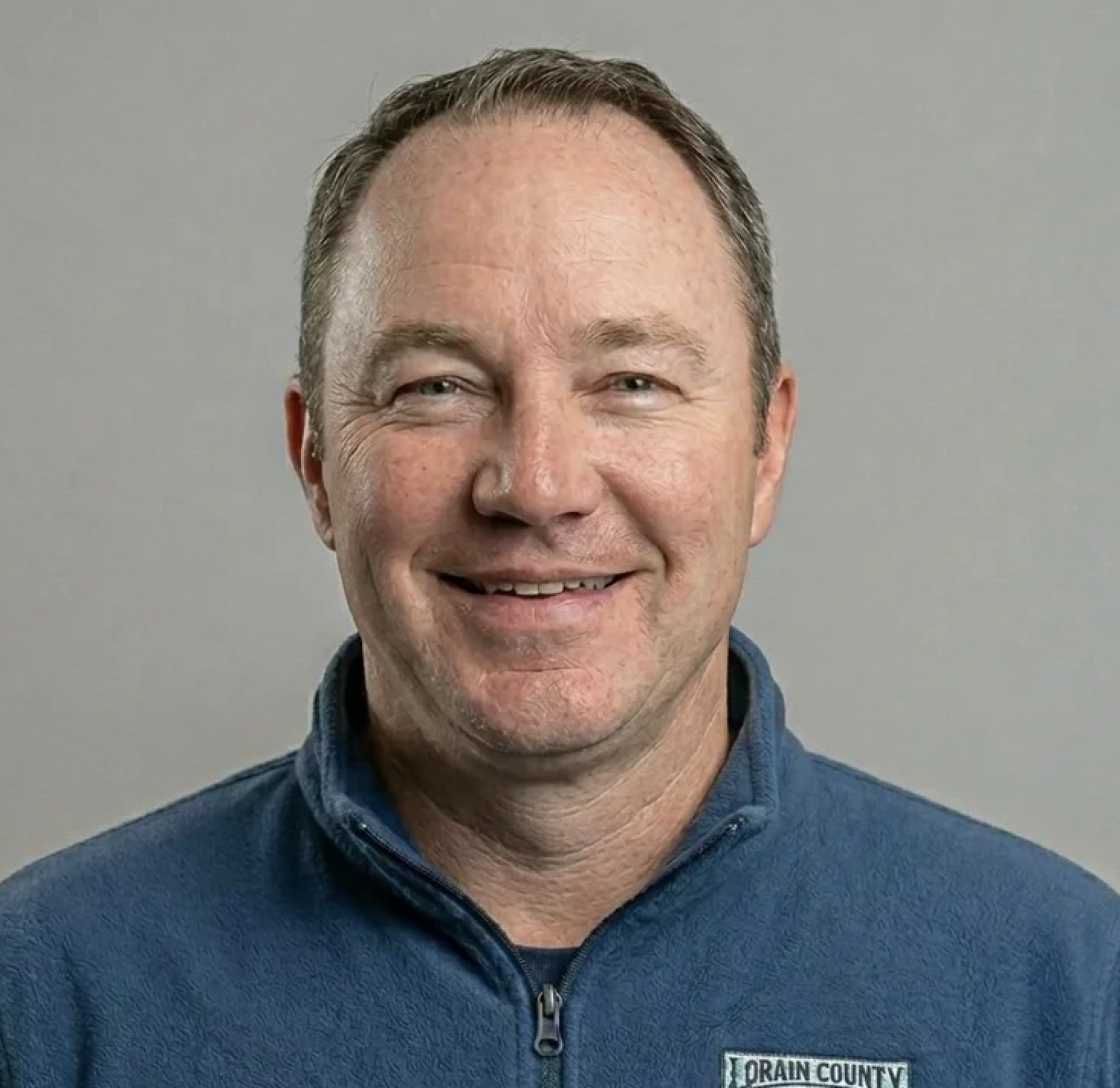 A man in a dark suit and white shirt smiling against a plain gray background.
