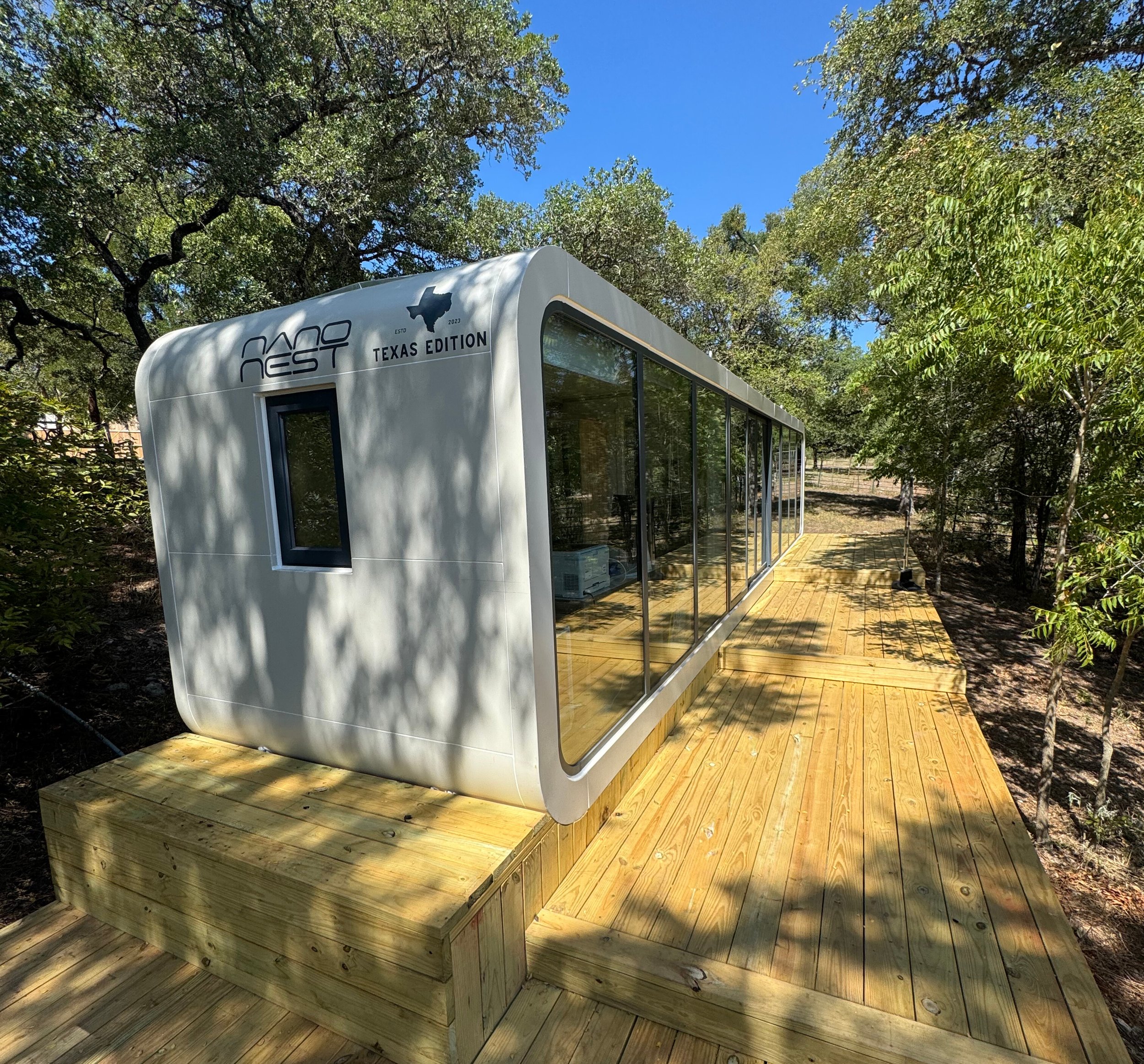 A modern tiny home with large glass windows and a rounded white exterior, labeled 'NAST Texas Edition' and 'ESTD 2023.' It is situated on a wooden deck in a wooded area under a clear blue sky.