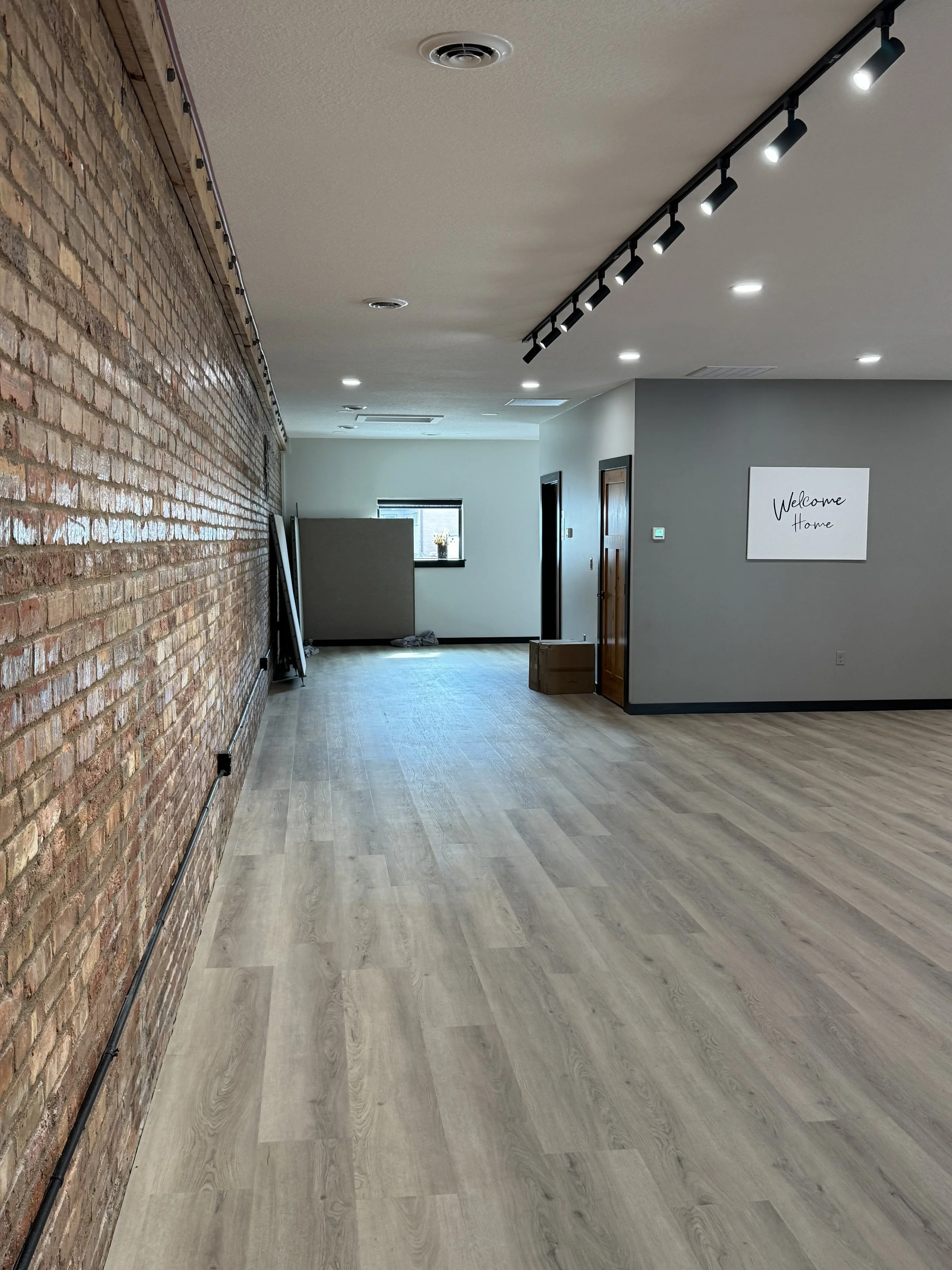 An interior room with a brick wall on the left and a white wall on the right. The ceiling has recessed lights and a track lighting system. There is a window at the back with a small ledge and a potted plant. A cardboard box is near a brown door on the right wall. A sign on the wall reads "Welcome Home." The floor is light wood laminate.