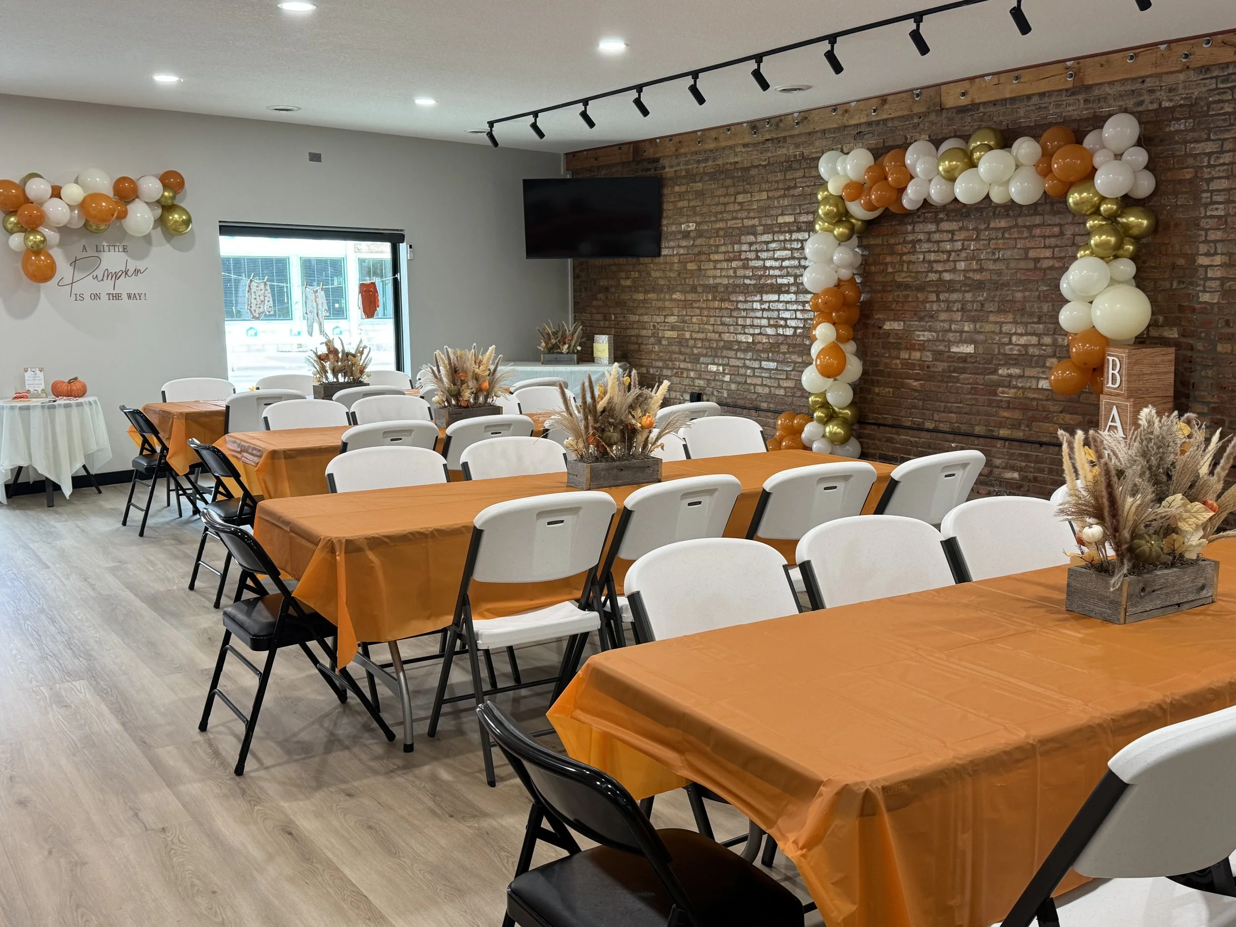 Indoor party setup with long tables covered in orange tablecloths, decorated with centerpieces of dried floral arrangements. A balloon arch in white, orange, and gold frames a brick wall backdrop. White and black chairs are arranged around the tables, and there is a small round table with pumpkins on the left. The room has wood flooring, track lighting, and a window with decorative hanging items outside.