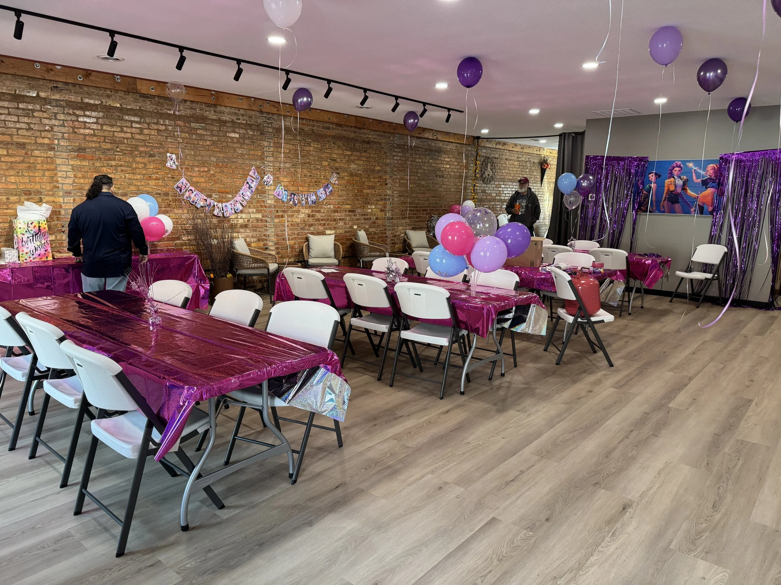 Decorated party room with pink and purple balloons, balloons on tables, pink tablecloths, and a banner on the wall that reads 'Happy Birthday'. There are chairs around the tables, some with cups and plates, and a few people preparing for the celebration.