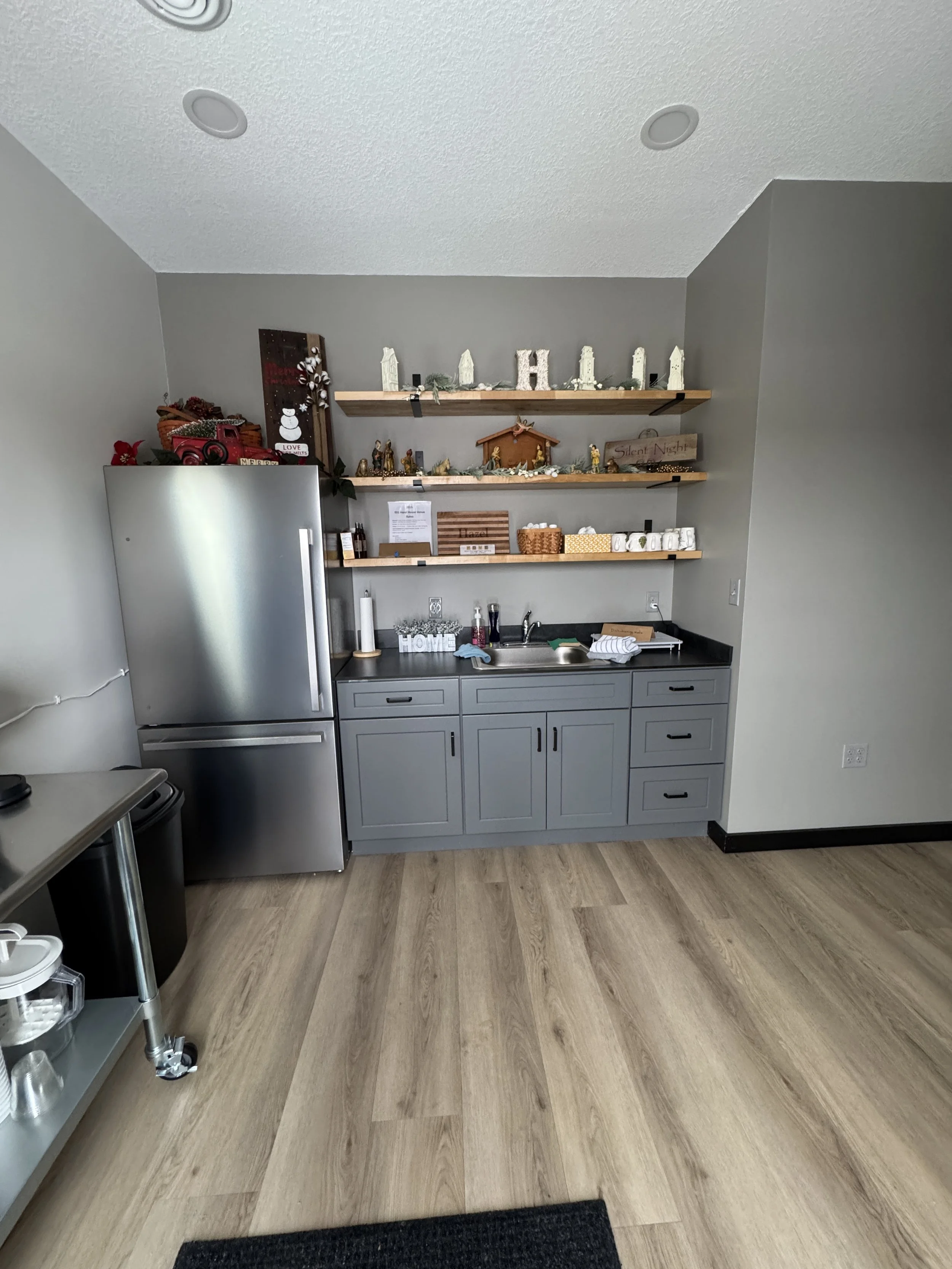 A modern kitchen area with gray cabinets, a stainless steel refrigerator, and wooden floating shelves decorated with Christmas items and nativity figurines.