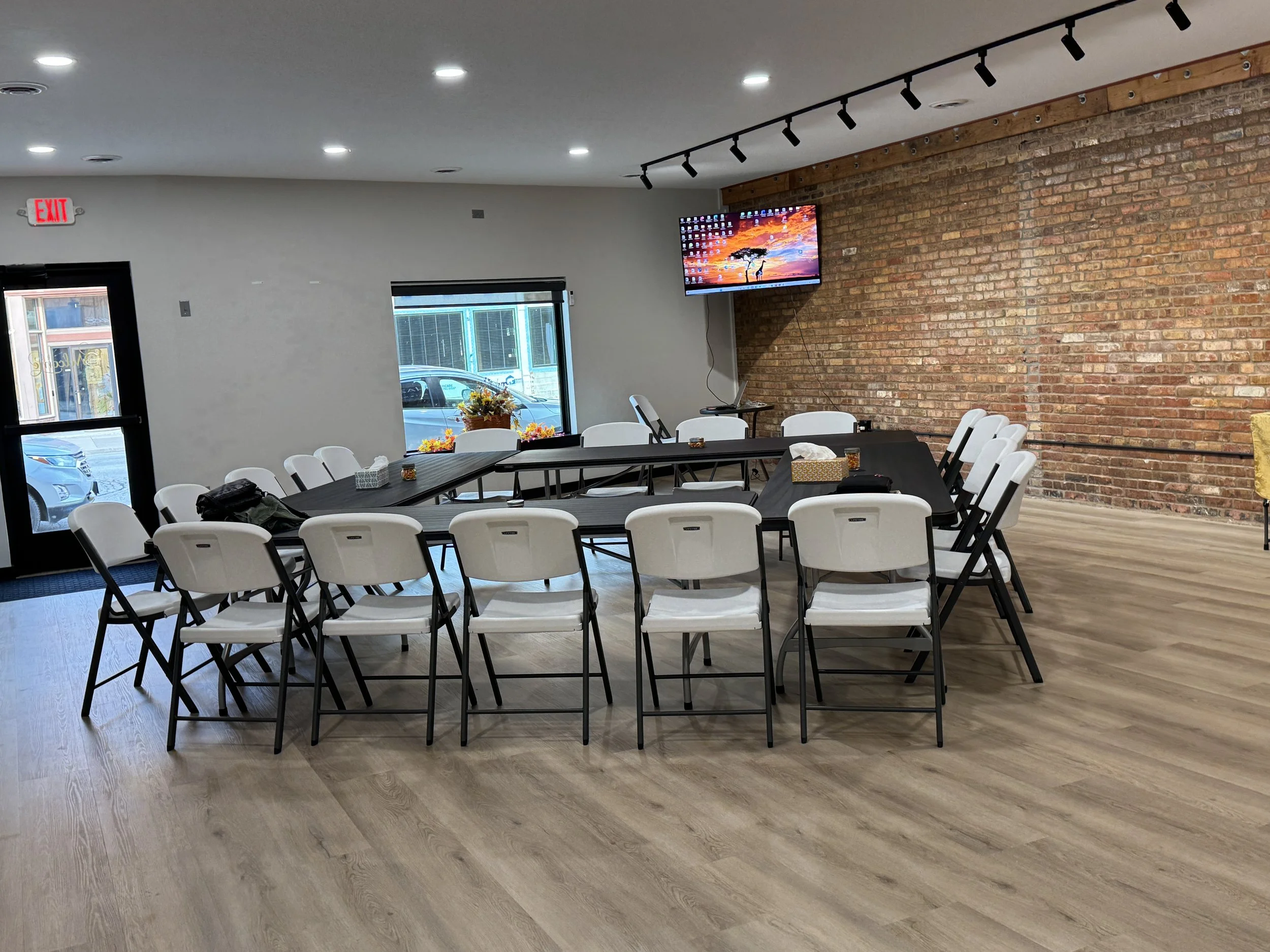 Meeting room with white chairs arranged around a U-shaped black conference table, a brick accent wall, a mounted television, and windows with view of parked cars outside.