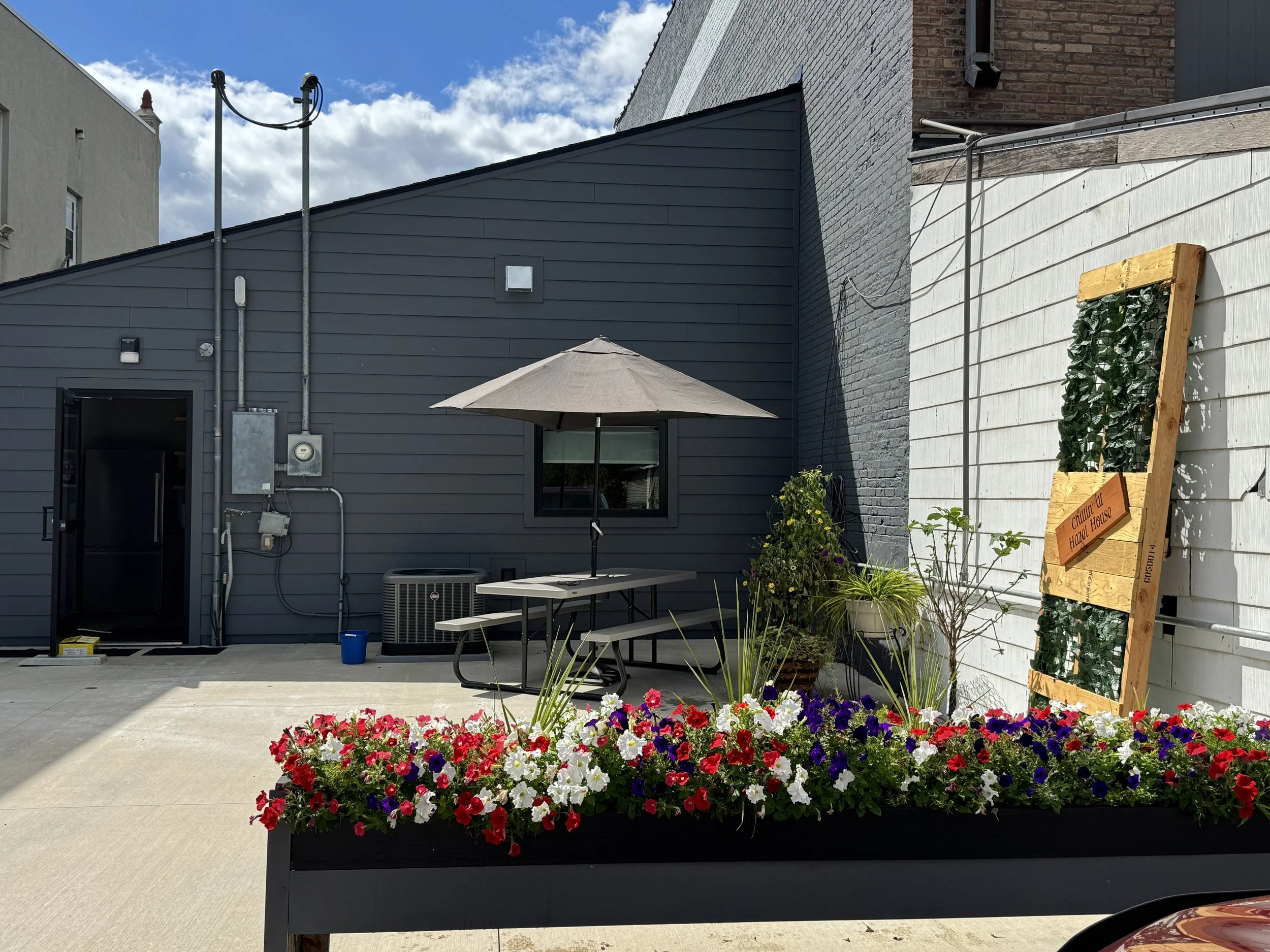 Urban outdoor patio with a flower bed, patio table with umbrella, plants, and a building with a blue and white wall.