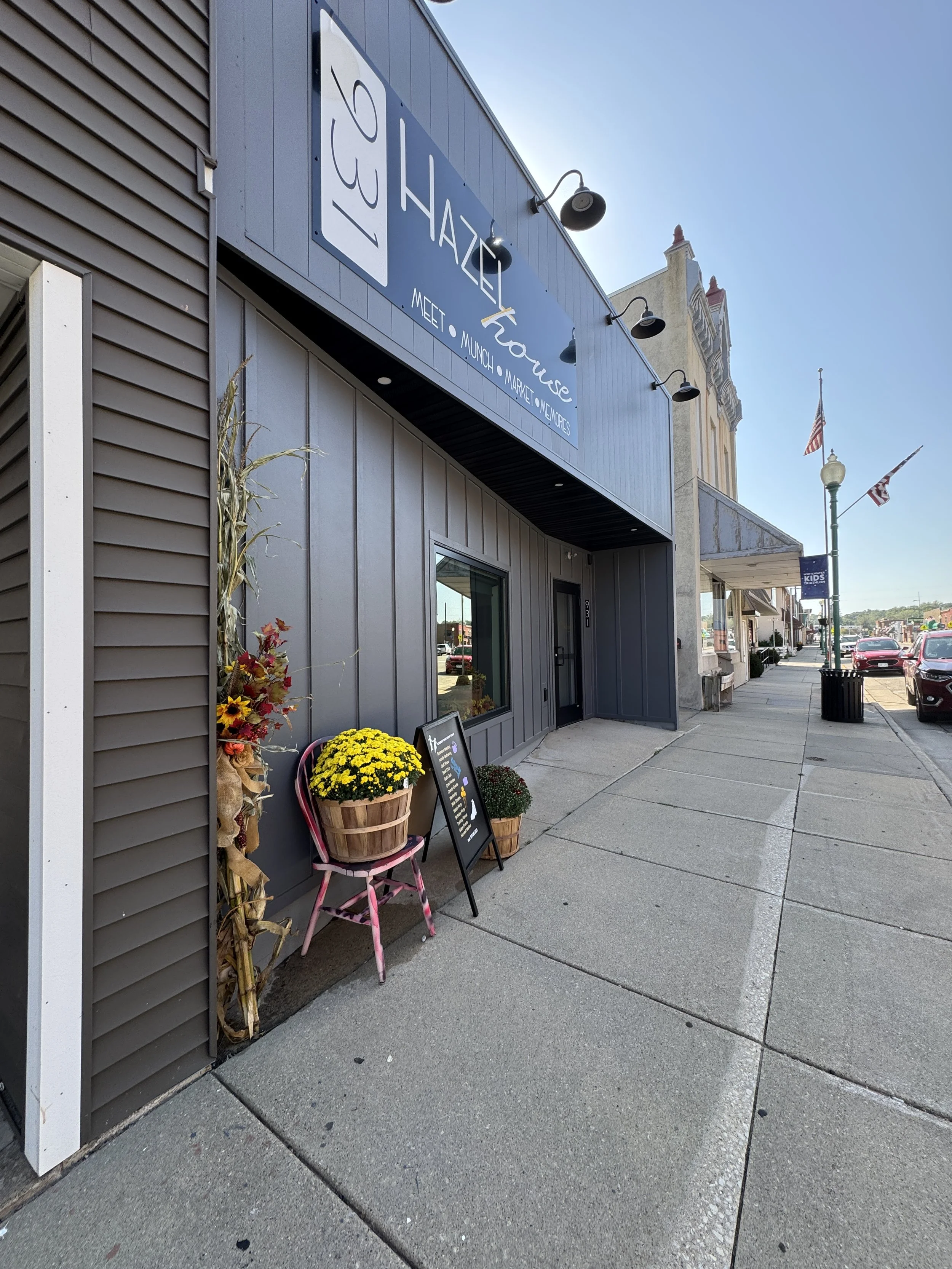 Exterior view of Hazel House, a shop with a gray facade and a large sign. Outside the shop, there are a pink chair, yellow potted flowers, and a blackboard sign. The sidewalk and street with parked cars are visible.