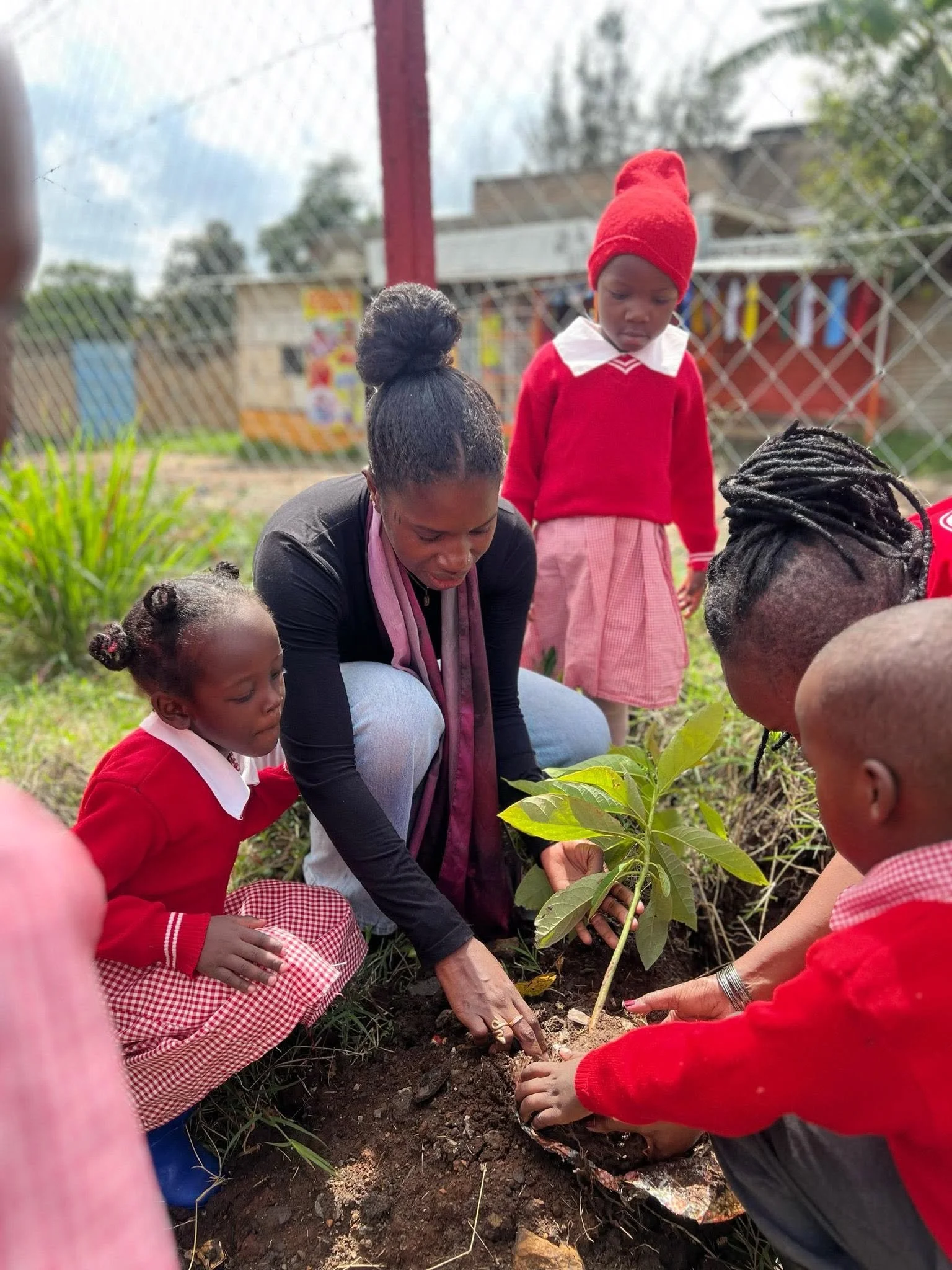 A group of children and an adult planting a small tree in the soil outdoors, with some children wearing red and pink school uniforms and an adult helping guide the planting.